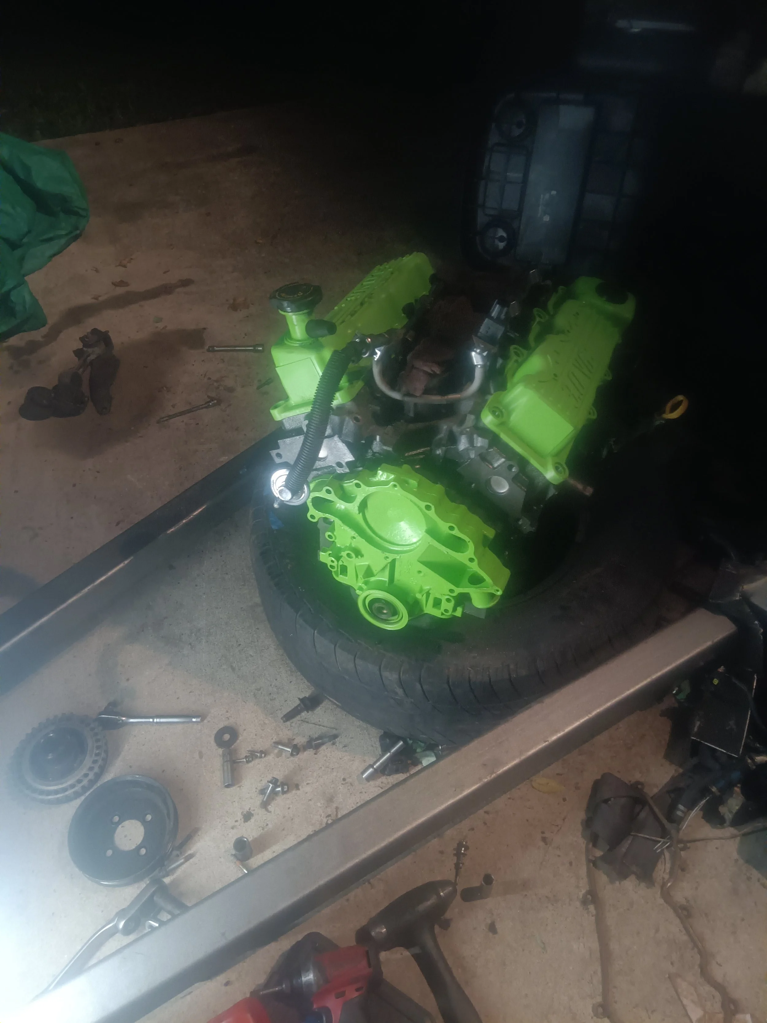 A car engine with bright green components placed on top of a tire, surrounded by tools and parts on a workbench in a garage.