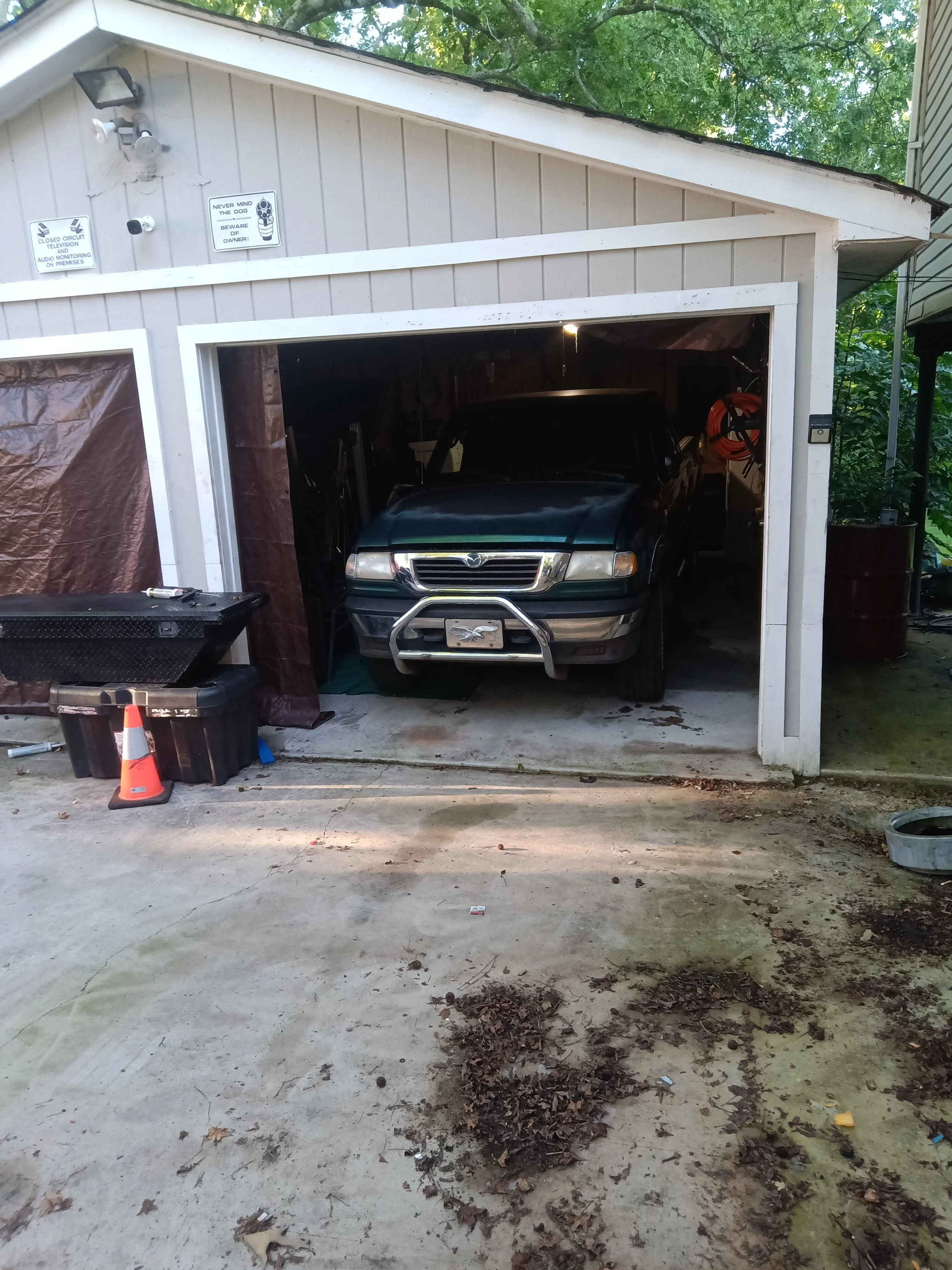 A black car parked inside a garage with a white exterior, brown curtains, and signs with warnings on the wall. The garage has tools and boxes inside, and there is a small traffic cone and leaf debris on the driveway outside.