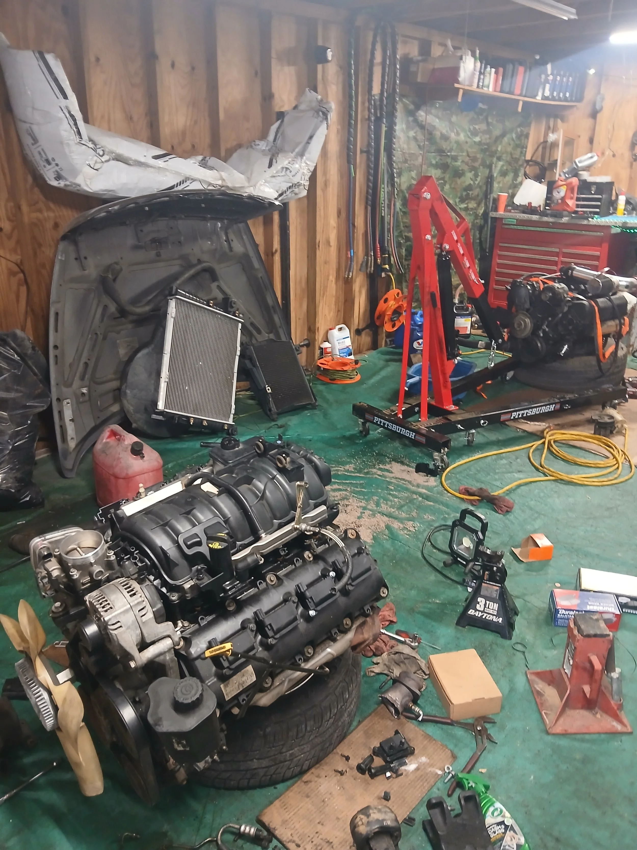 Garage with car engine on a tire, various tools, car parts, and equipment scattered around, including a radiator, oil, lift, and shelves with containers and books.