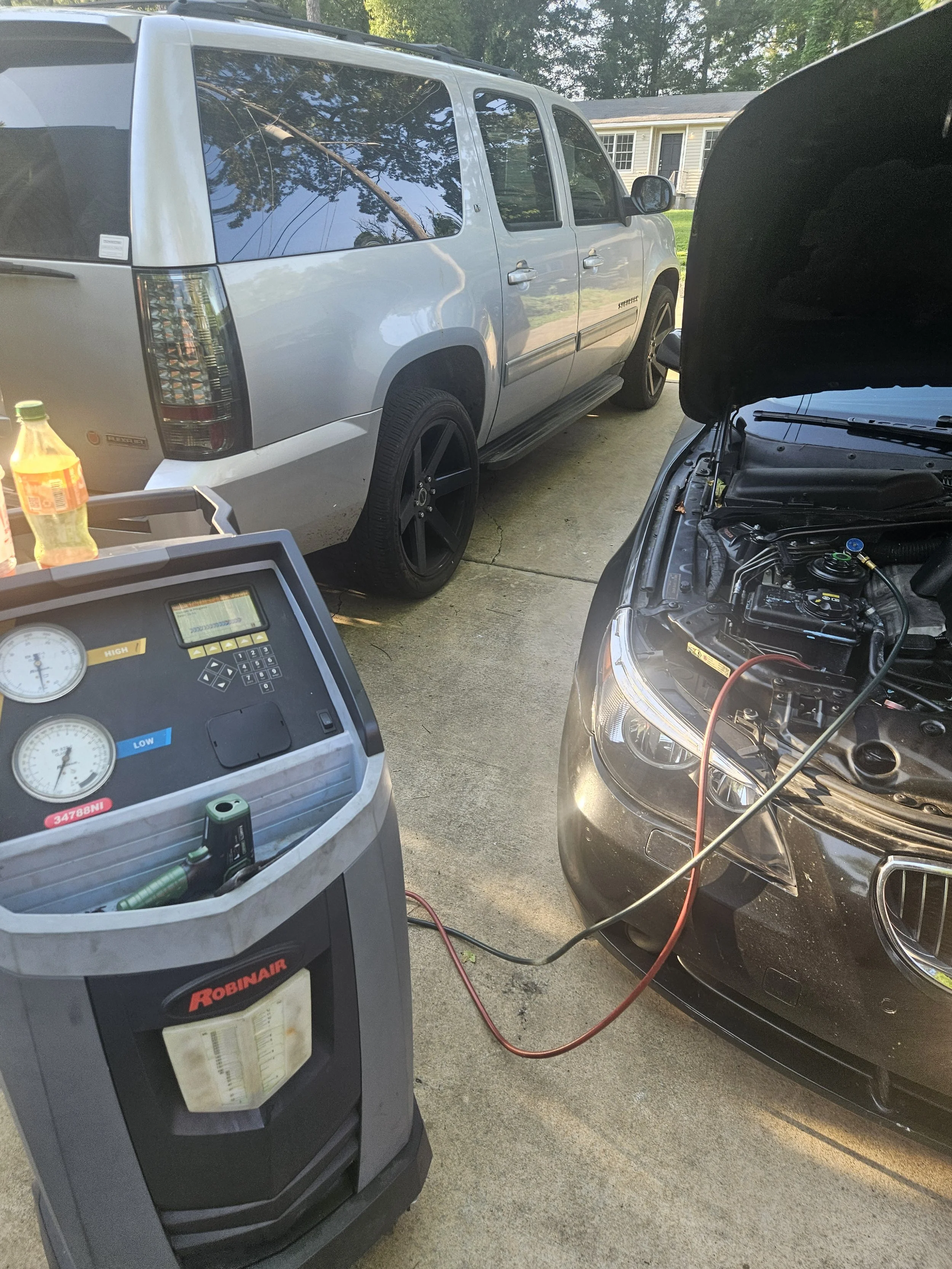 Car undergoing air conditioning service with hood open, connected to a Robinair A/C service machine, in a driveway with another silver SUV parked nearby.