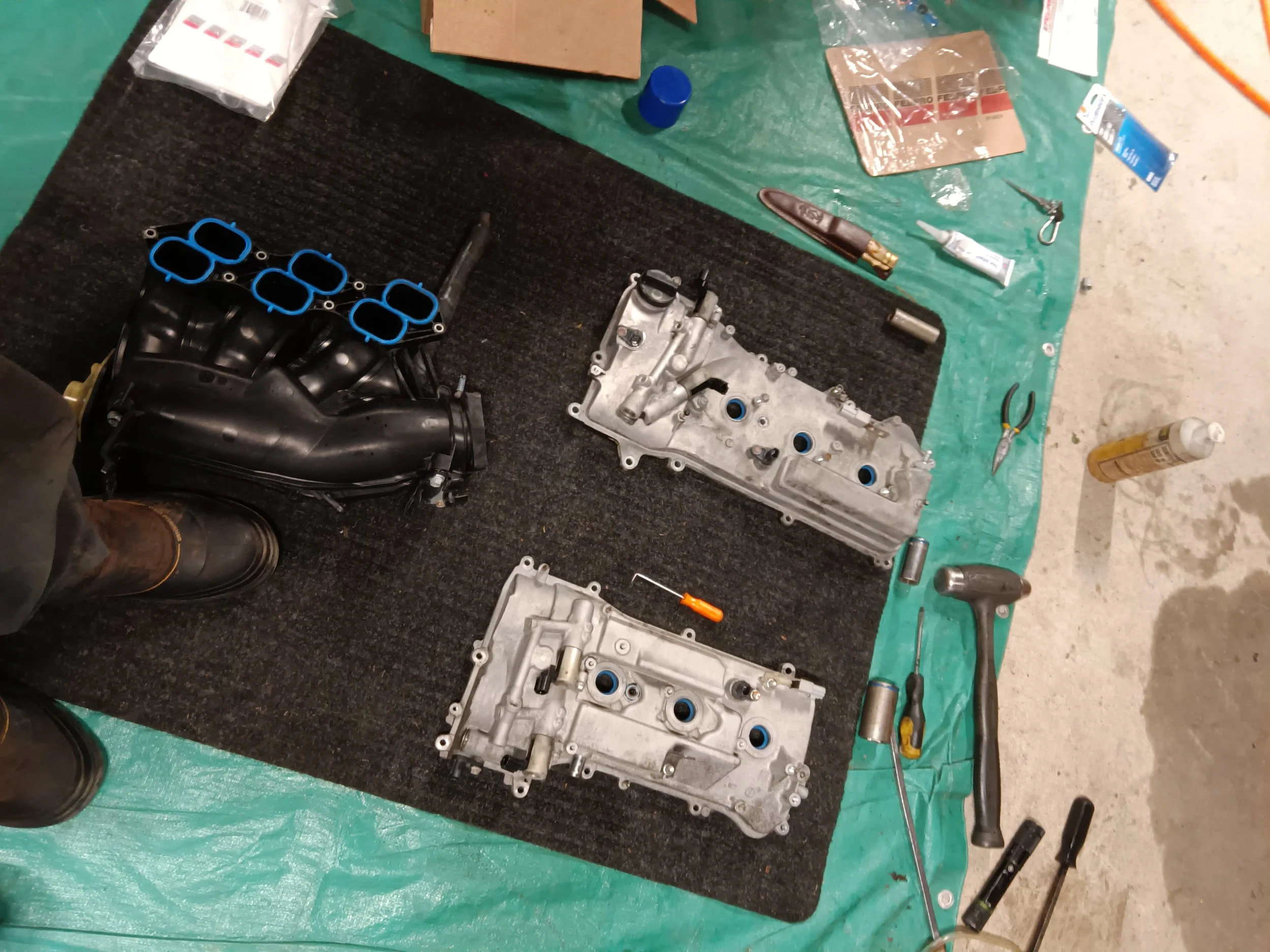 Disassembled car engine components on a workbench with tools, including intake manifold, valve covers, screwdriver, hammer, pliers, and lubricant bottle.