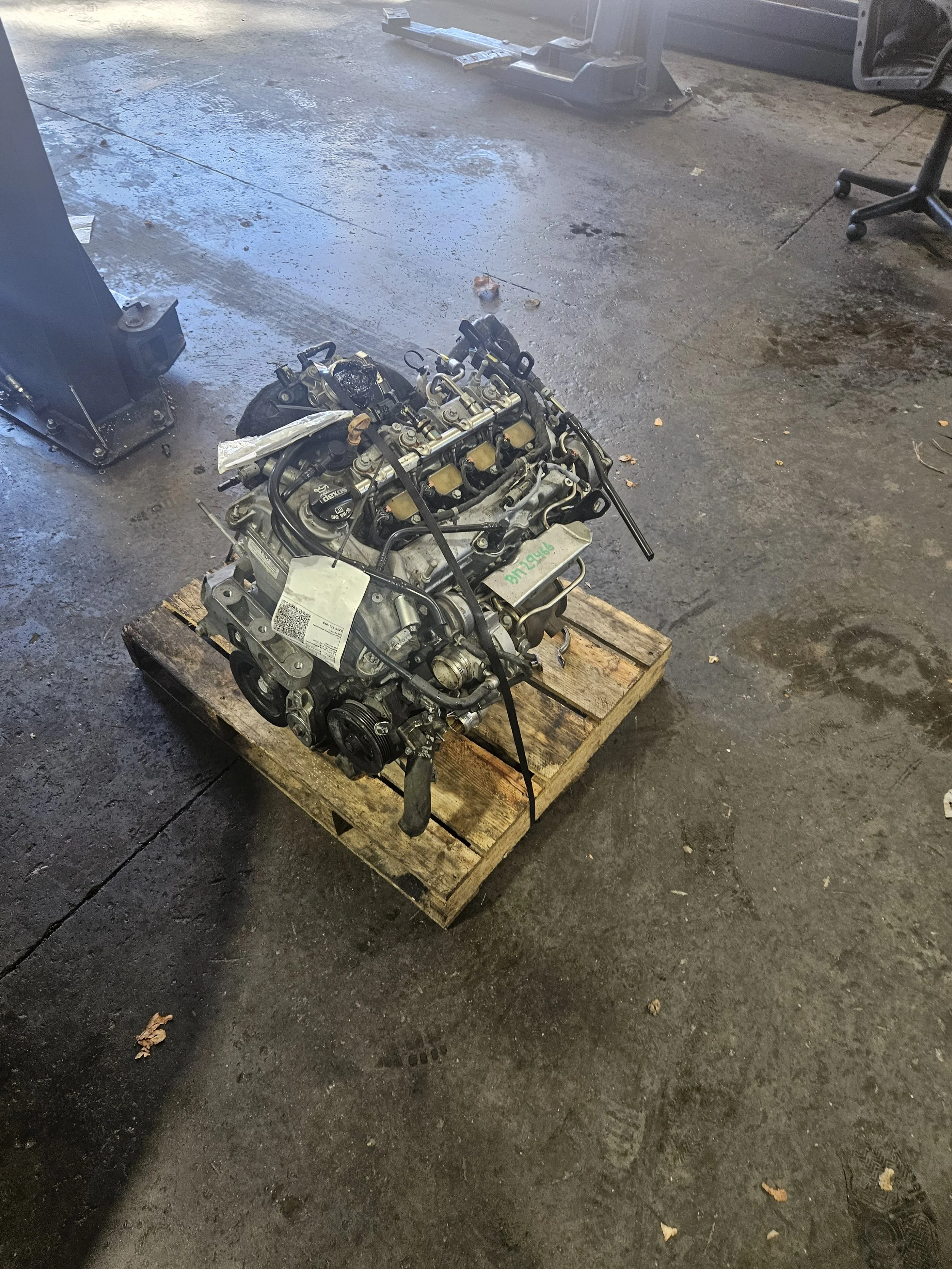 Auto engine placed on a wooden pallet in an industrial workshop with concrete floor and some equipment in the background.
