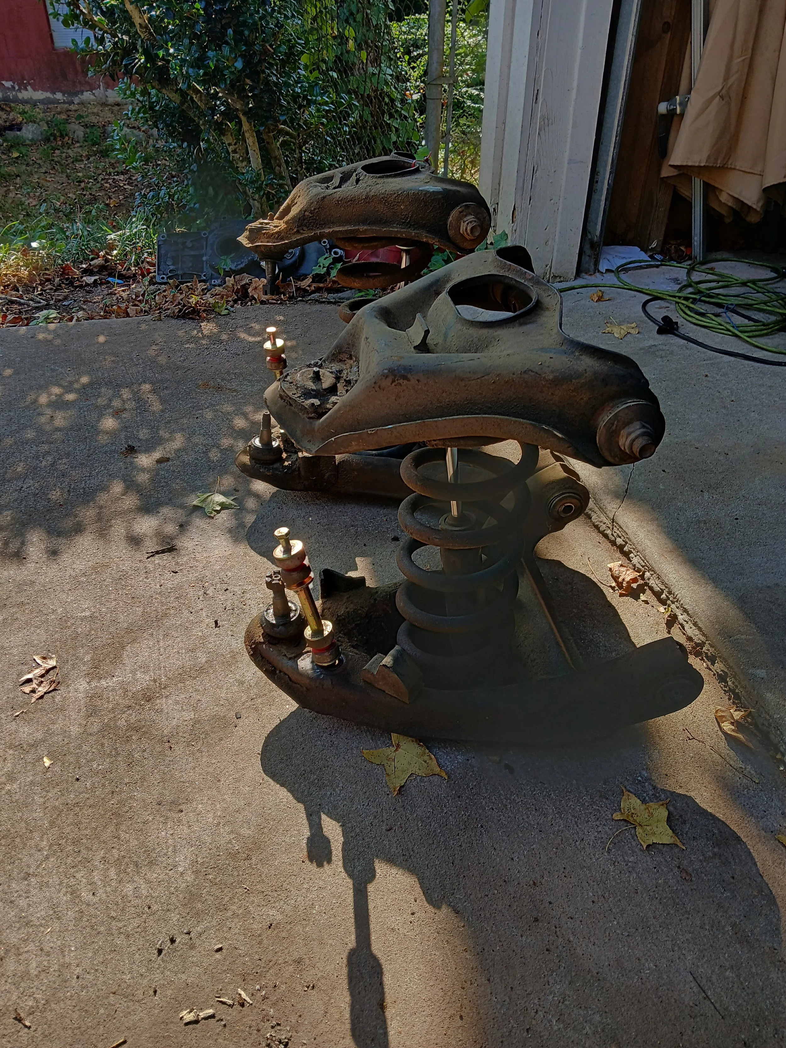 Disassembled car suspension components, including control arms and springs, placed on a concrete driveway with leaves and tree shadows around.