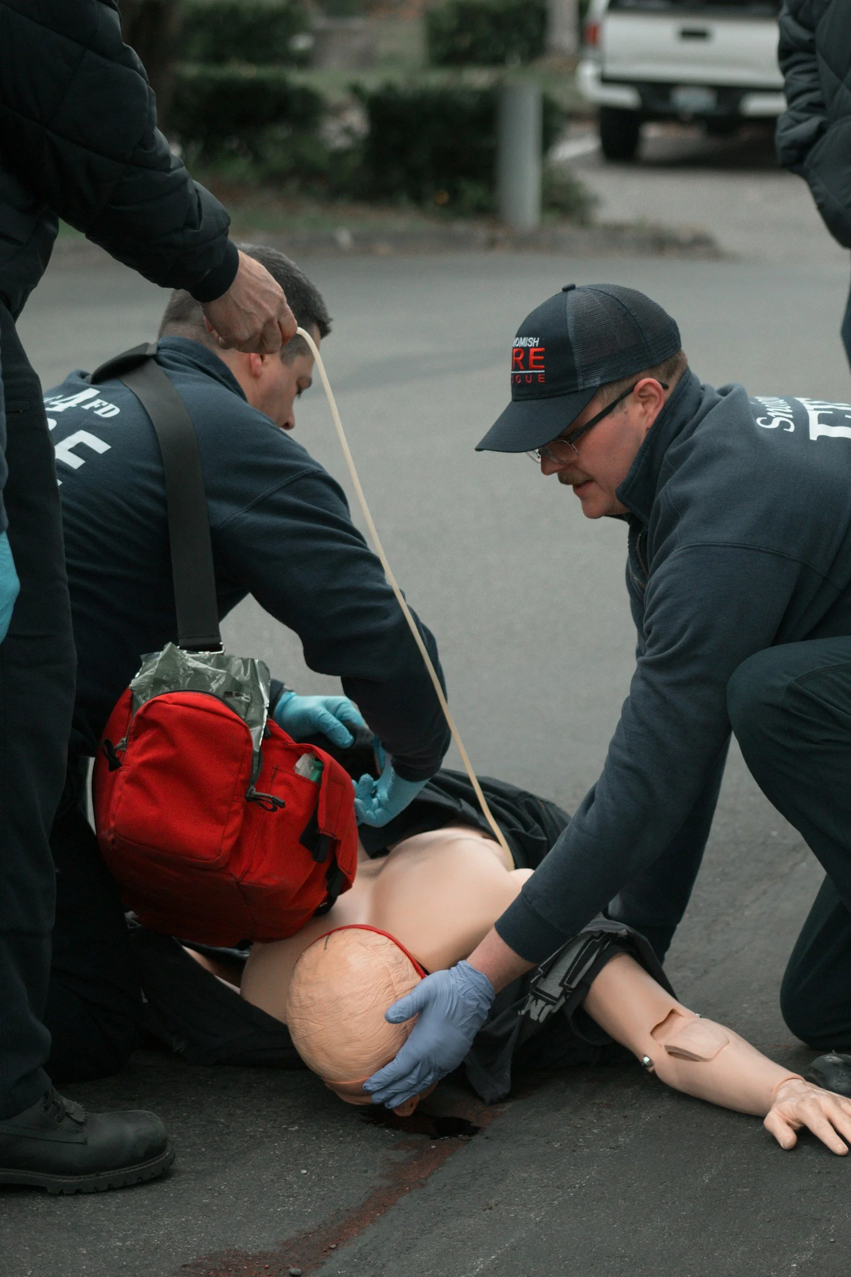 Emergency medical responders attending to a person lying on the road, simulating CPR on a training dummy during a practice drill.