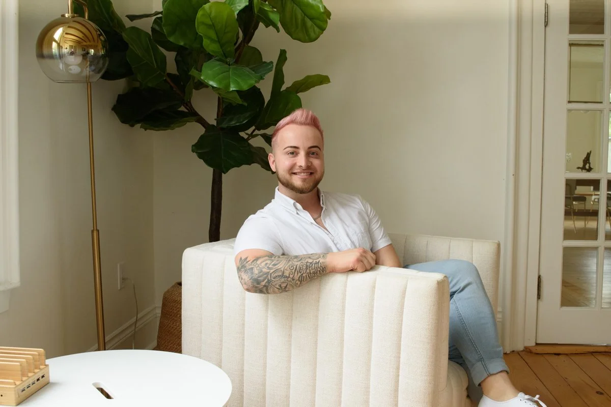 A man with pink hair and tattoos, wearing a white shirt and light blue jeans, sitting on a striped white and beige armchair in a living room with a large green plant, a gold floor lamp, and a glass-paneled door.