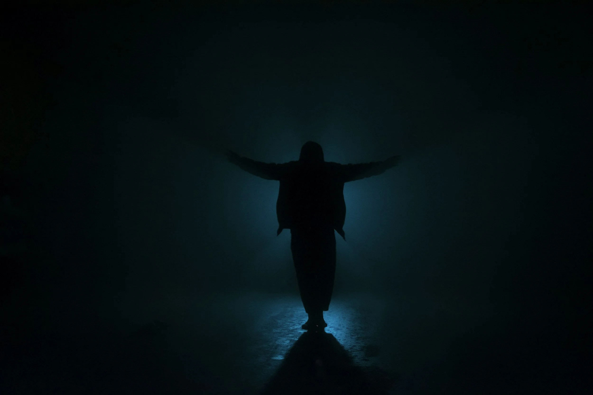 Silhouette of a person with arms outstretched, standing on a surface illuminated by a blue light, with a dark background.