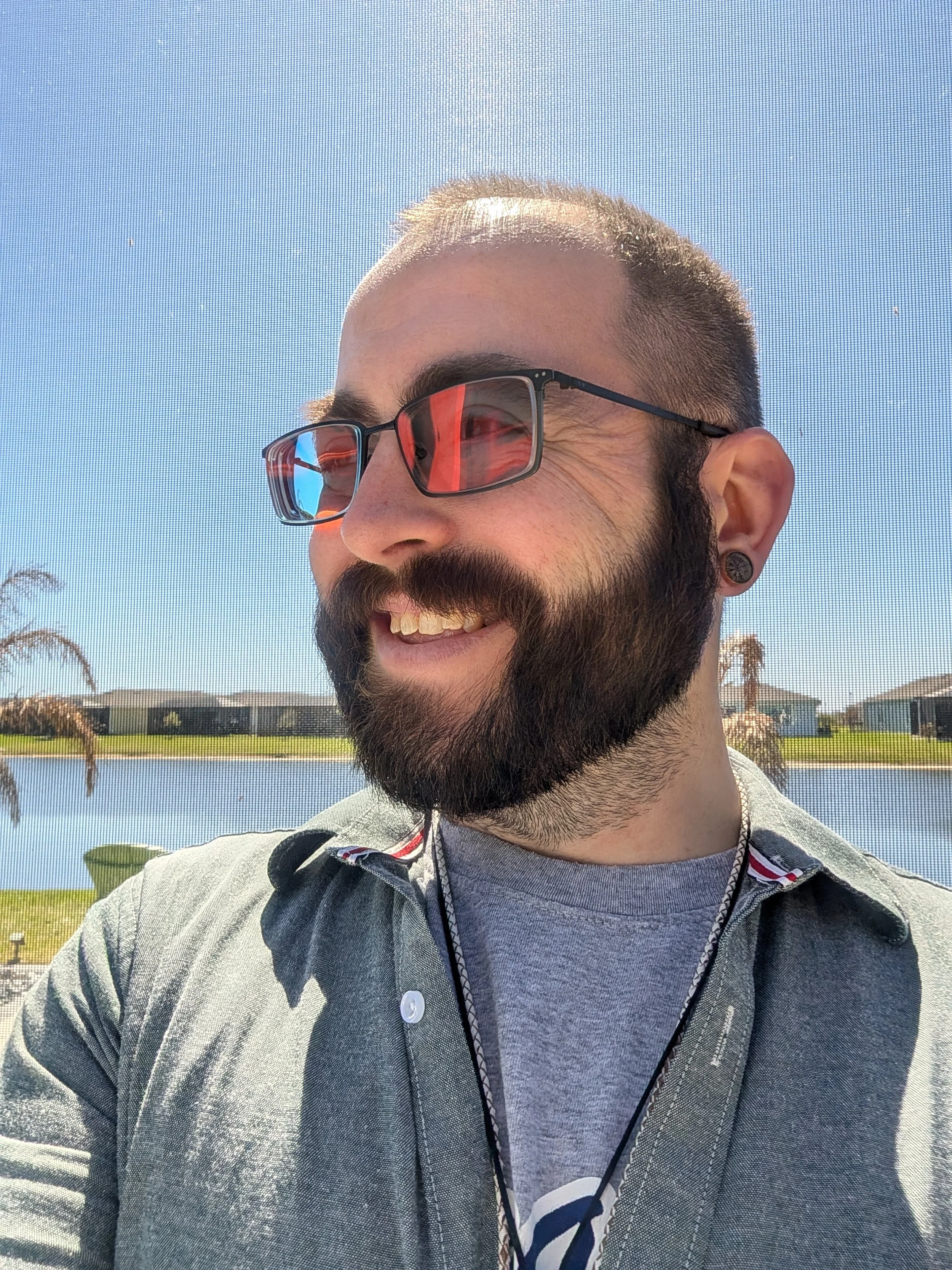 A man with a beard and sunglasses smiling outdoors near a lake, with houses in the background on a sunny day.
