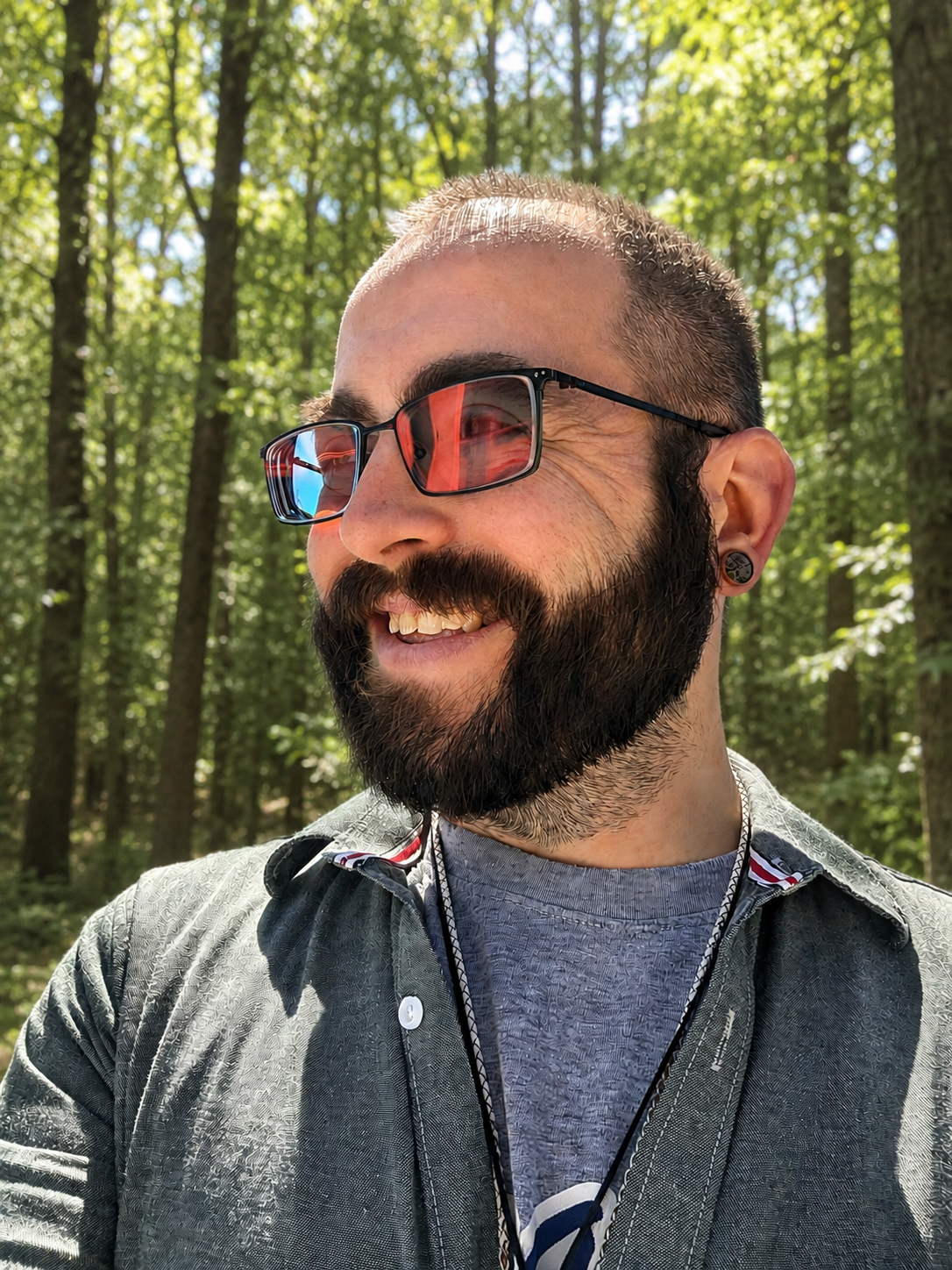 Close-up of a smiling man with sunglasses, a beard, and earrings, outdoors in a wooded area with sunlight filtering through trees.
