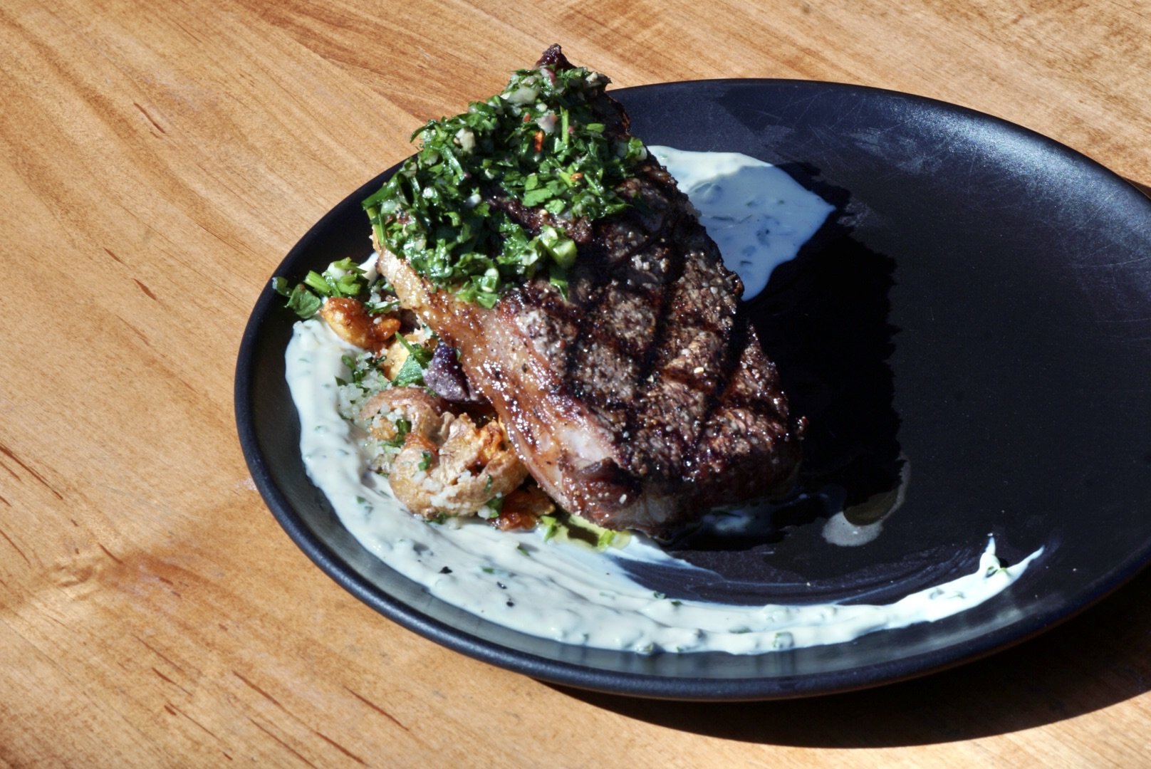 Grilled beef steak topped with chopped herbs, served on a black plate with creamy sauce, garnished with fried onions, on a wooden table.