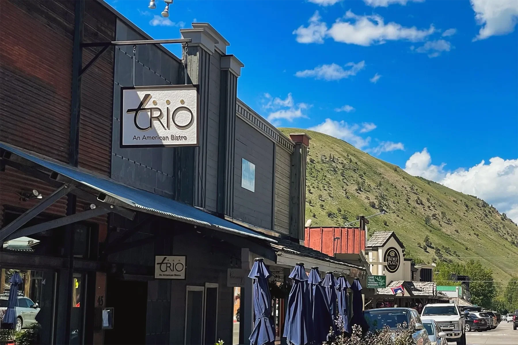 Exterior view of a restaurant named 'Trio: An American Bistro,' with outdoor seating and umbrellas, located in a mountain town under a blue sky with clouds.