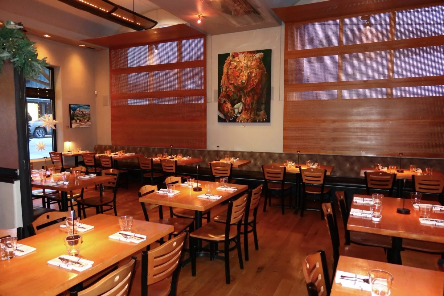 Empty restaurant with wooden tables and chairs, set with glasses, utensils, and napkins, warm lighting, bear artwork on wall, window with decorations.