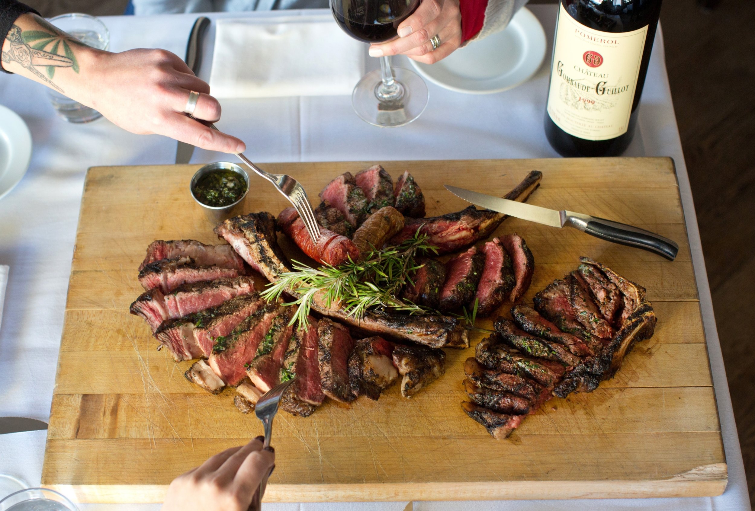 Sliced grilled steaks on a wooden cutting board garnished with herbs, a person with a tattooed arm holding a fork, wine glass, all on a white tablecloth.