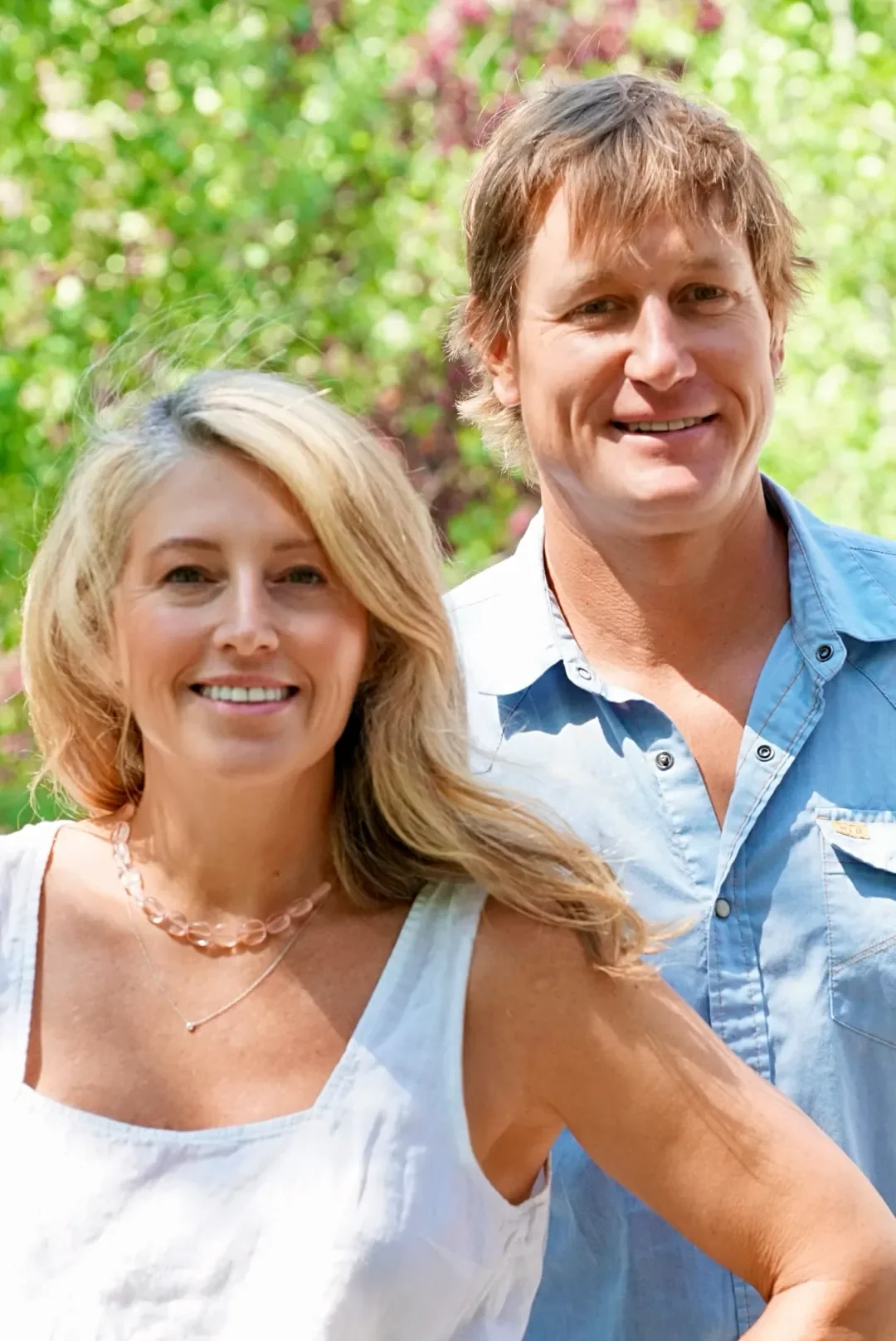 A smiling woman with blonde hair wearing a white sleeveless top and a necklace, standing next to a smiling man with light brown hair, wearing a light blue button-up shirt, outdoors with green and pink foliage in the background.