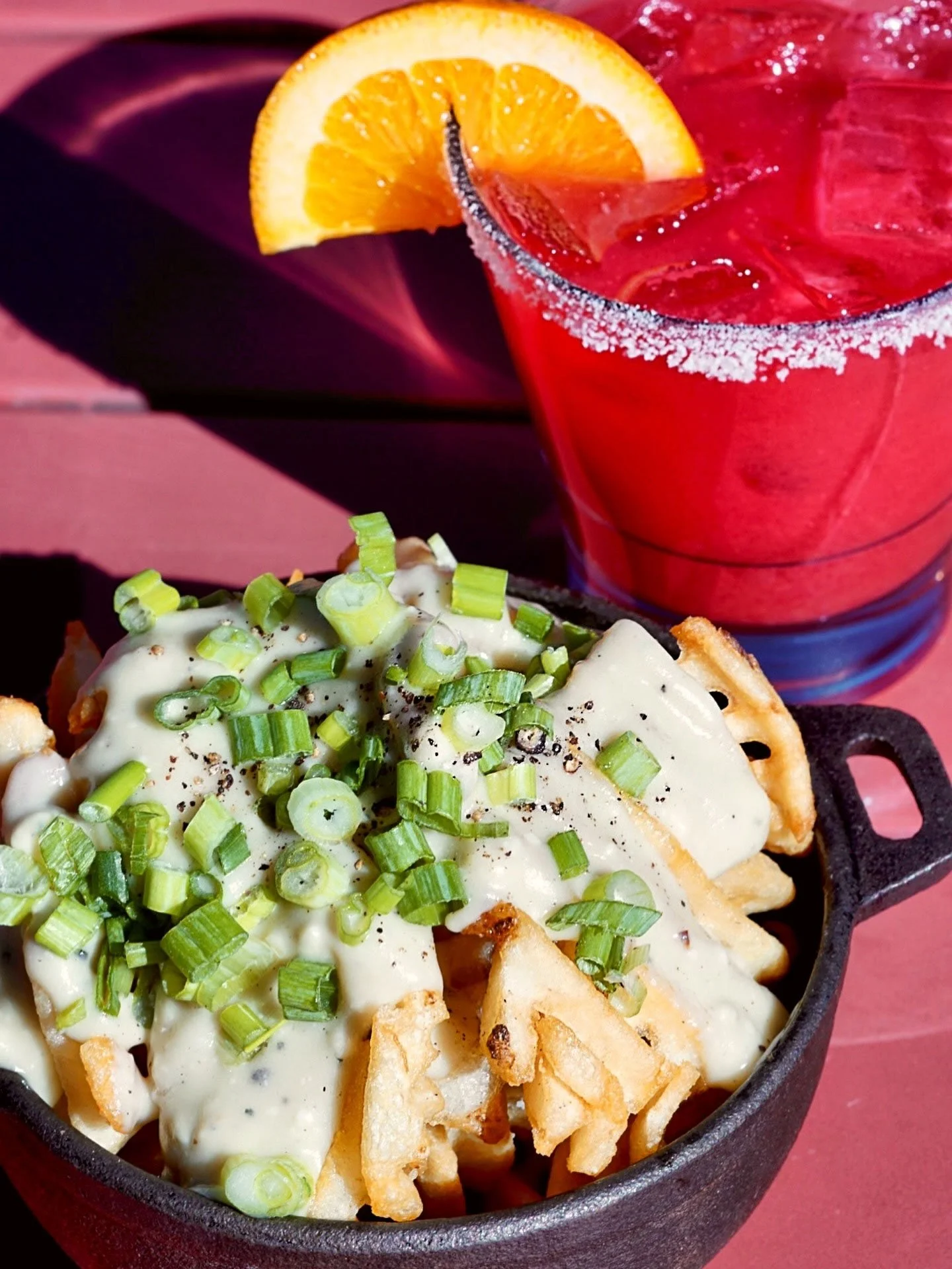 Close-up of a baked dish topped with chopped green onions and creamy sauce, served in a small cast iron skillet, with a side of potato chips and a thick red drink garnished with orange slice.