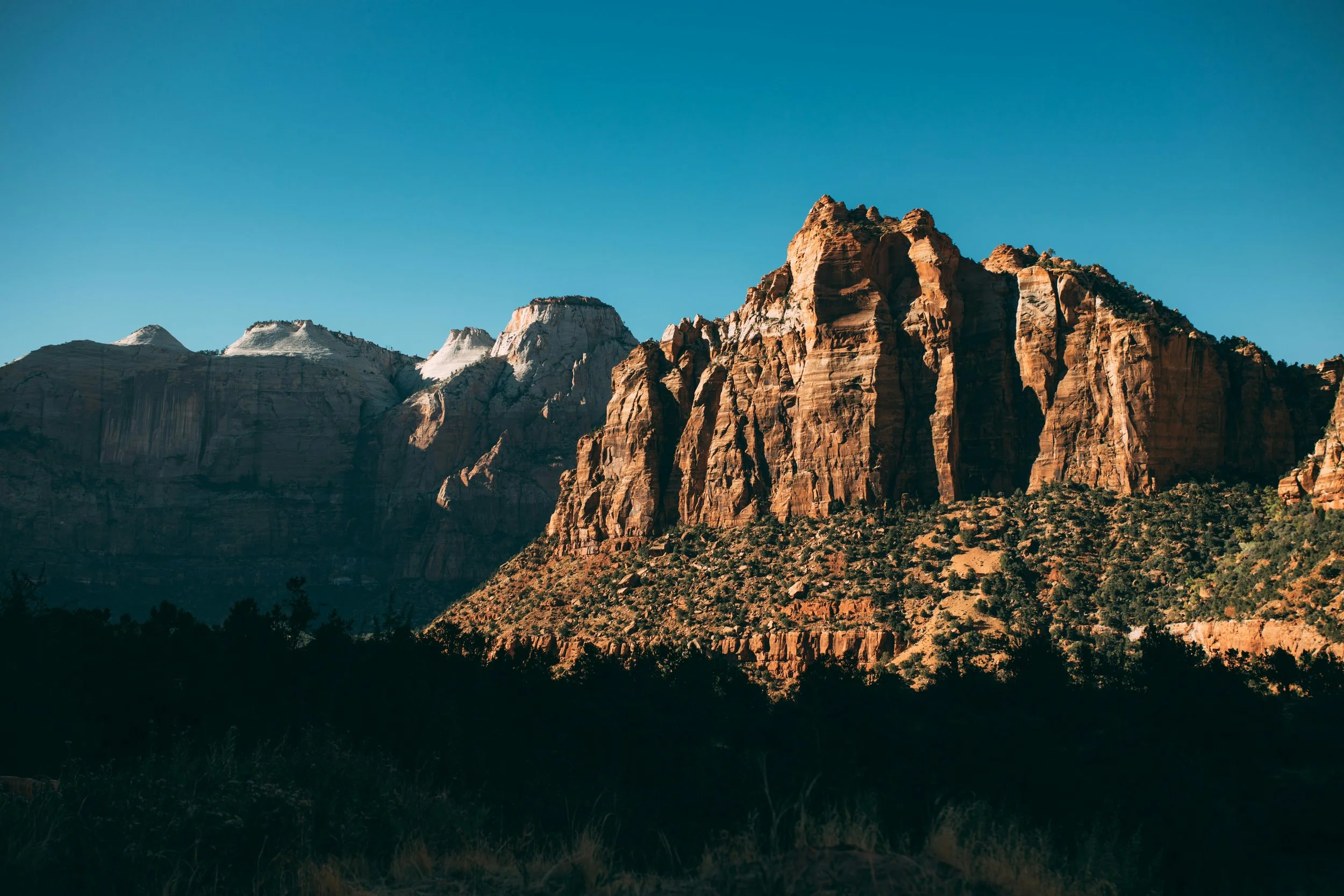 A mountain range with rugged cliffs and peaks under a clear blue sky, with some snow visible on the higher elevations and dark green trees at the base.