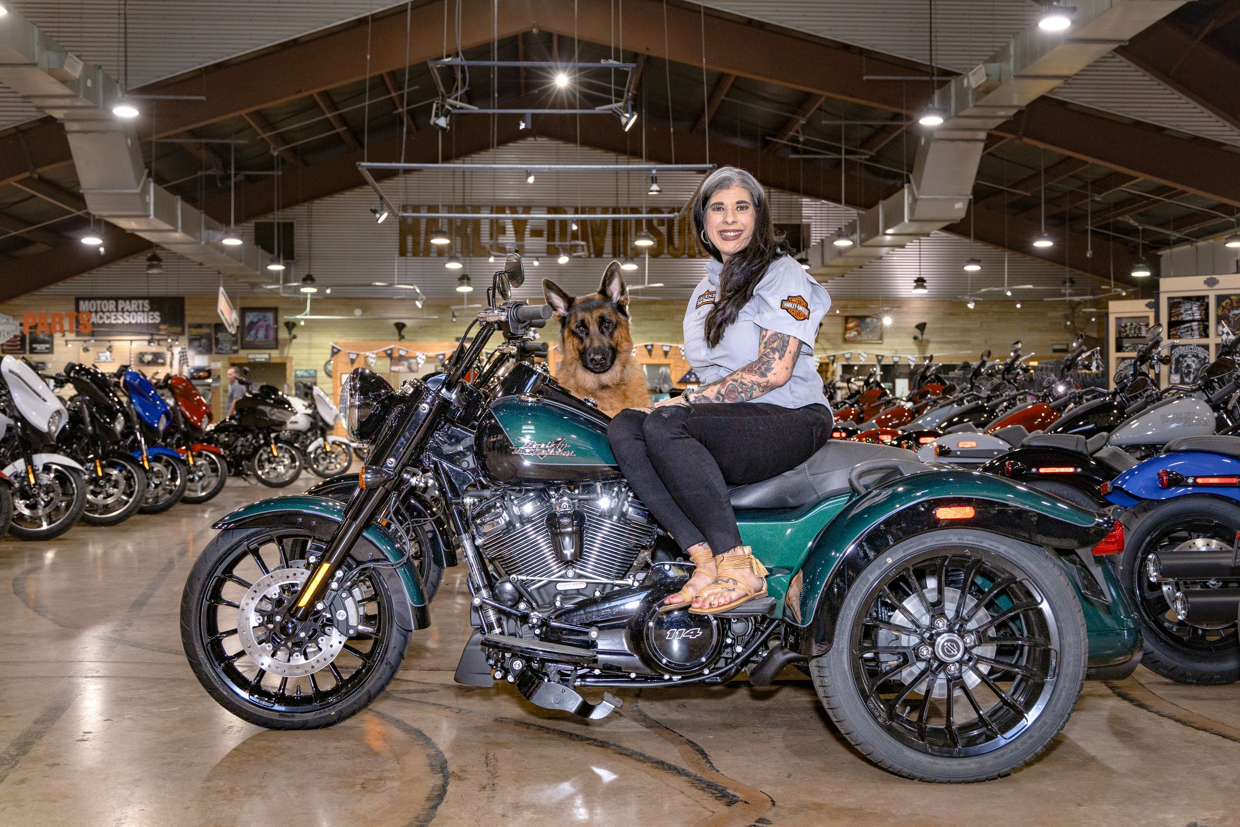 A woman with dark hair, tattoos, and a smile, sitting on a green motorcycle inside a motorcycle dealership, with a German shepherd dog beside her. The dealership has multiple motorcycles on display and is well-lit.