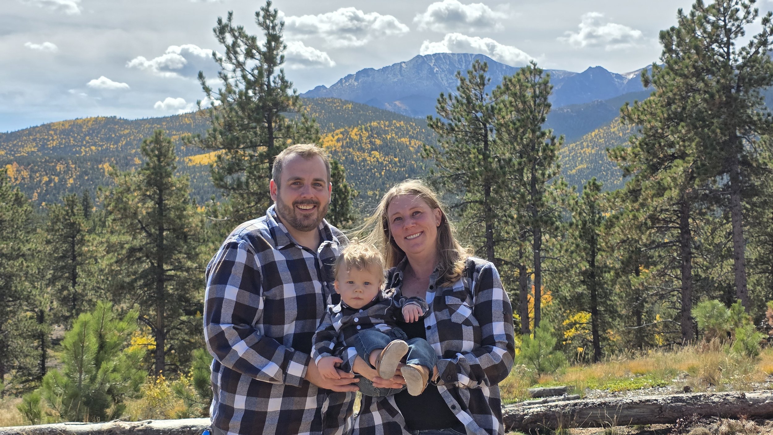 Dr. Greer, Kyle Kemble, and their son, standing outdoors in a forested mountainous area with pine trees and mountains in the background. They are smiling and wearing plaid shirts.