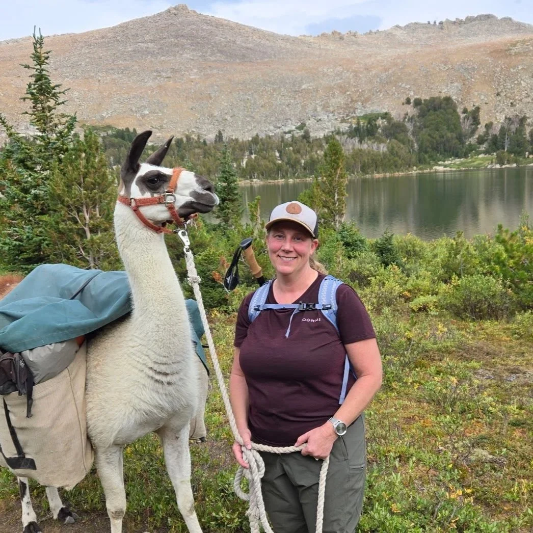 A woman standing outdoors near a lake with a llama carrying supplies; she is holding the llama's lead and smiling at the camera, surrounded by trees and mountains in the background.