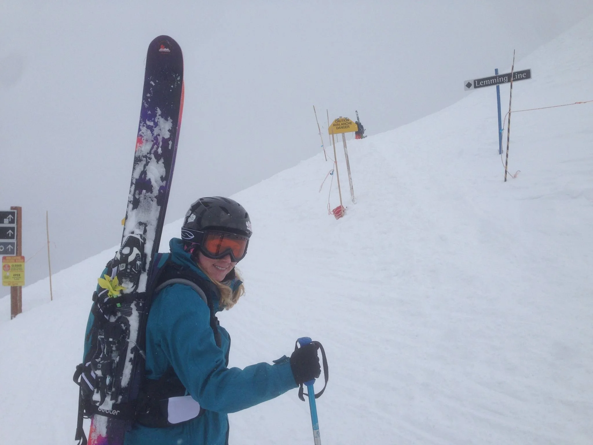 Dr. Greer dressed in winter gear with a helmet, goggles, and a blue jacket, standing in snowy mountain terrain with a snowboard attached to her backpack, holding a ski pole, smiling at the camera.