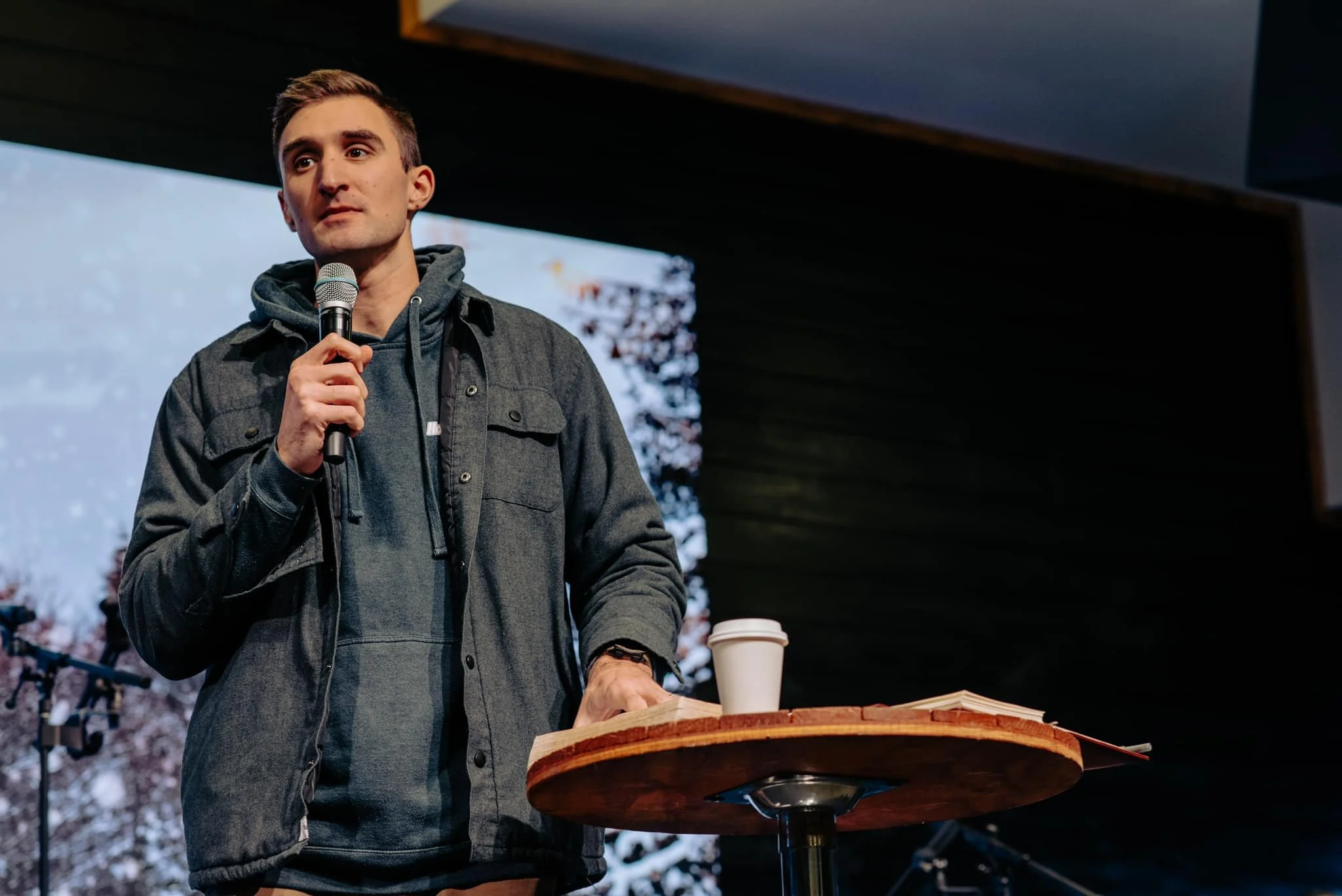 A young man holds a microphone while standing behind a wooden table with a paper coffee cup and an open book. He wears a hoodie and a jacket, and appears to be speaking or presenting in an indoor setting with a large screen or window showing trees in the background.