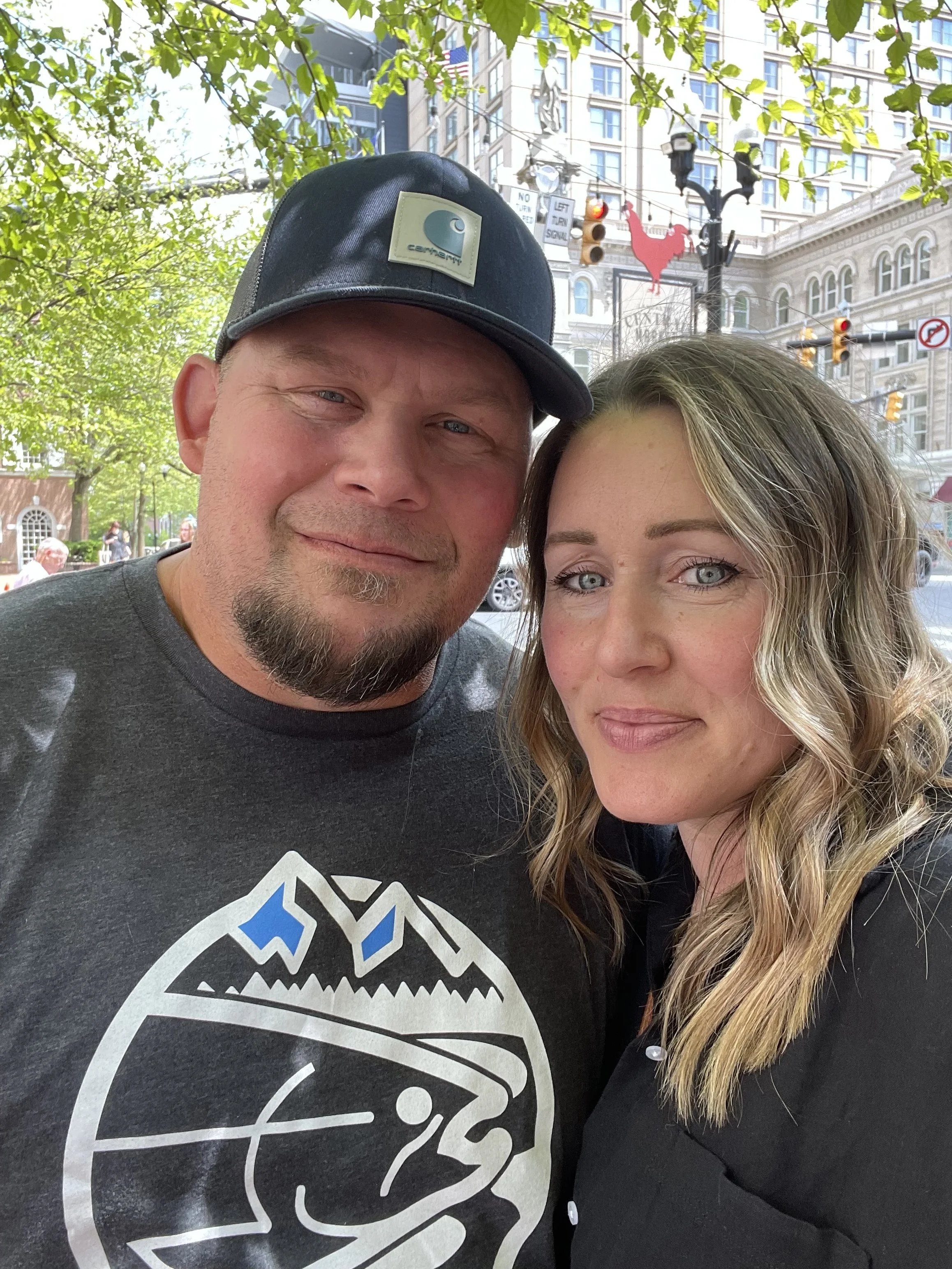 A man and woman taking a selfie outdoors on a city street with green trees, tall buildings, traffic lights, and street decorations visible in the background.