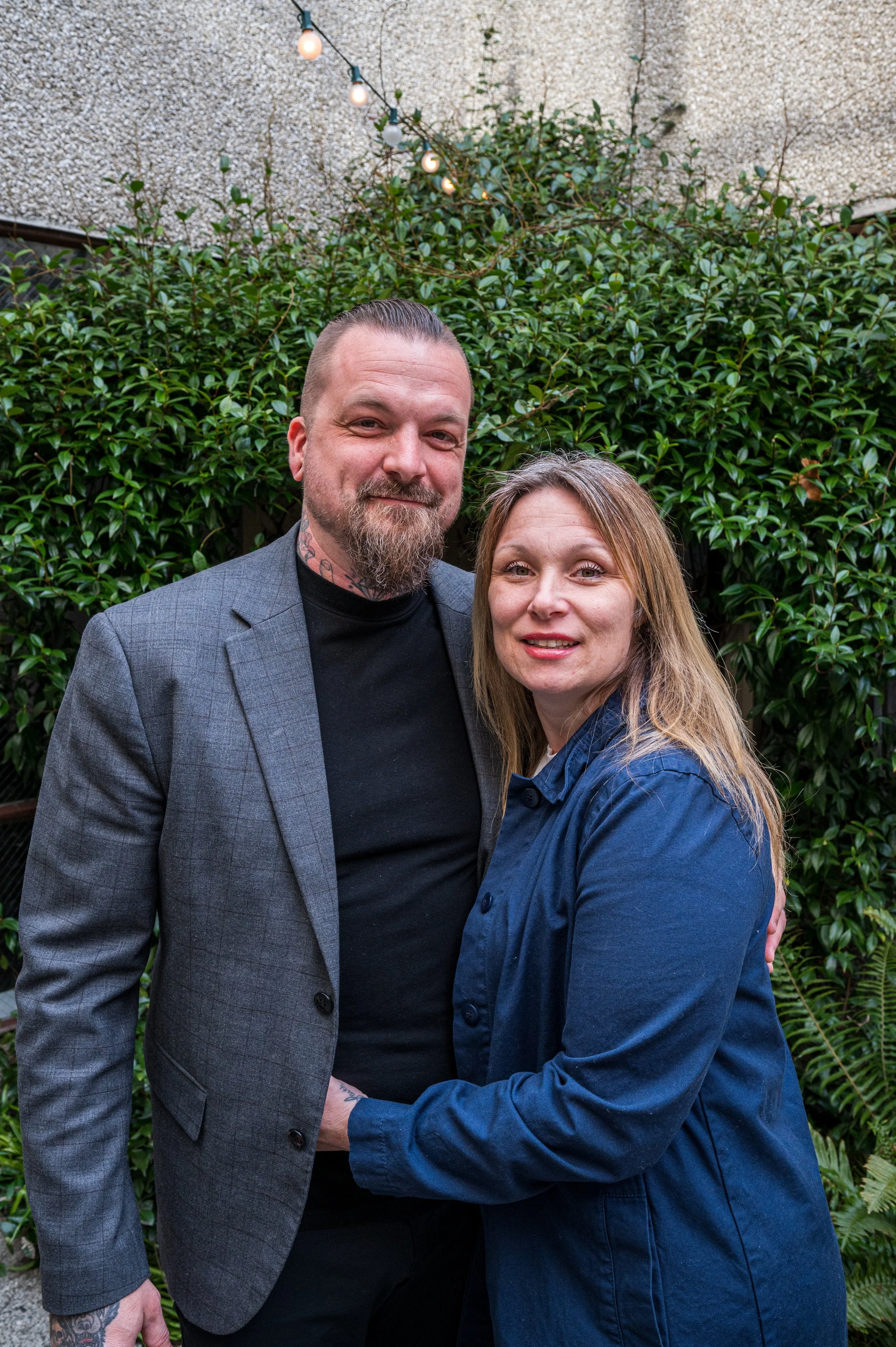 A man and woman standing close together outdoors near a large green leafy bush, smiling at the camera. The man has a beard and tattoos, wearing a gray blazer and black shirt. The woman has long, light brown hair, wearing a blue denim jacket.