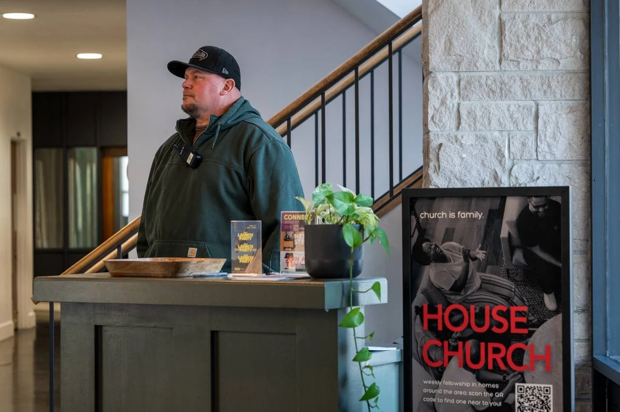 A man in a black hoodie and black cap standing at a reception desk inside a church or community center. There are pamphlets, a potted plant, and a sign with a QR code promoting house church nearby.