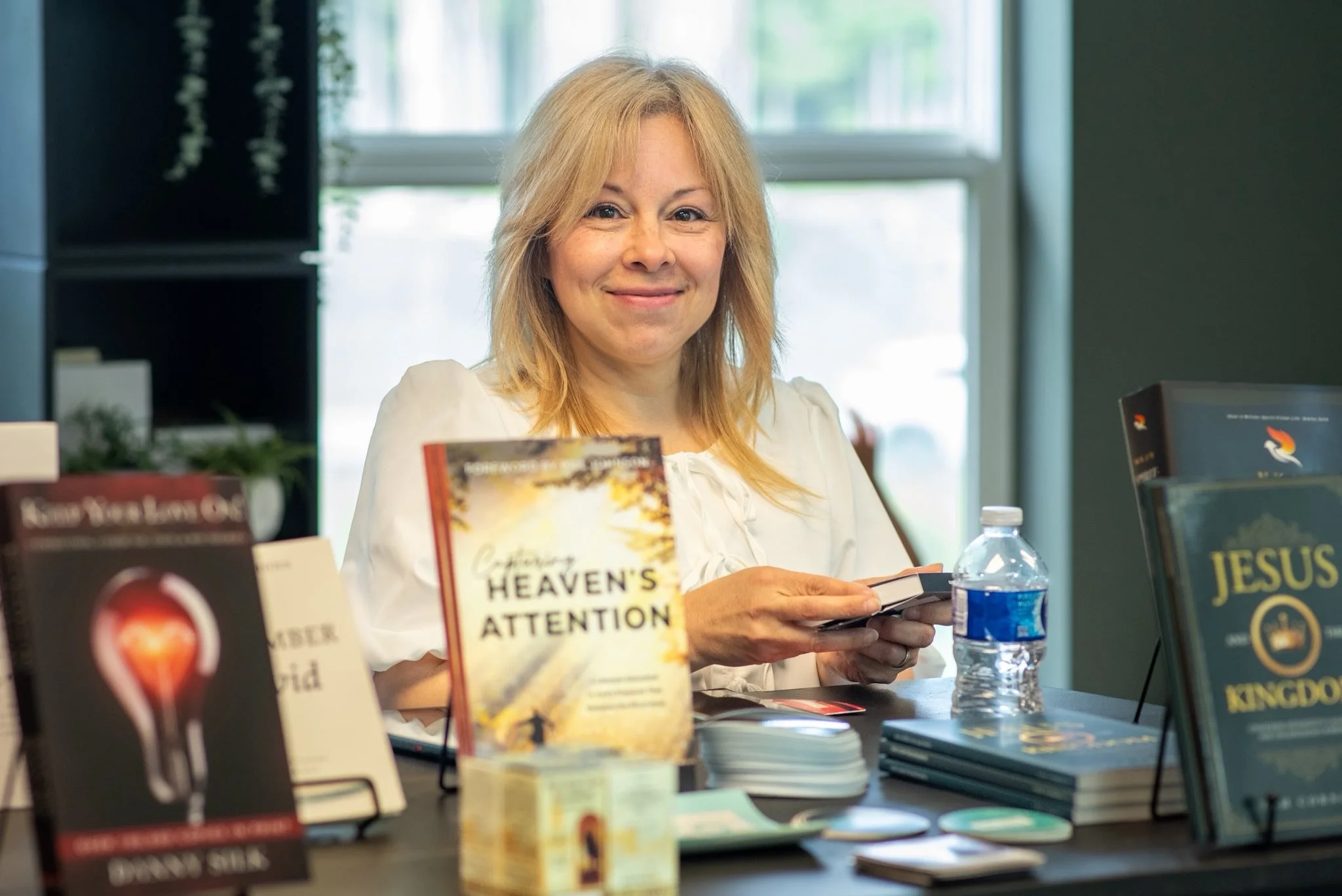 A woman with blonde hair smiling and holding a phone at a book signing or display event. Books about Christianity and Heaven are on the table in front of her, including 'Heaven's Attention'.
