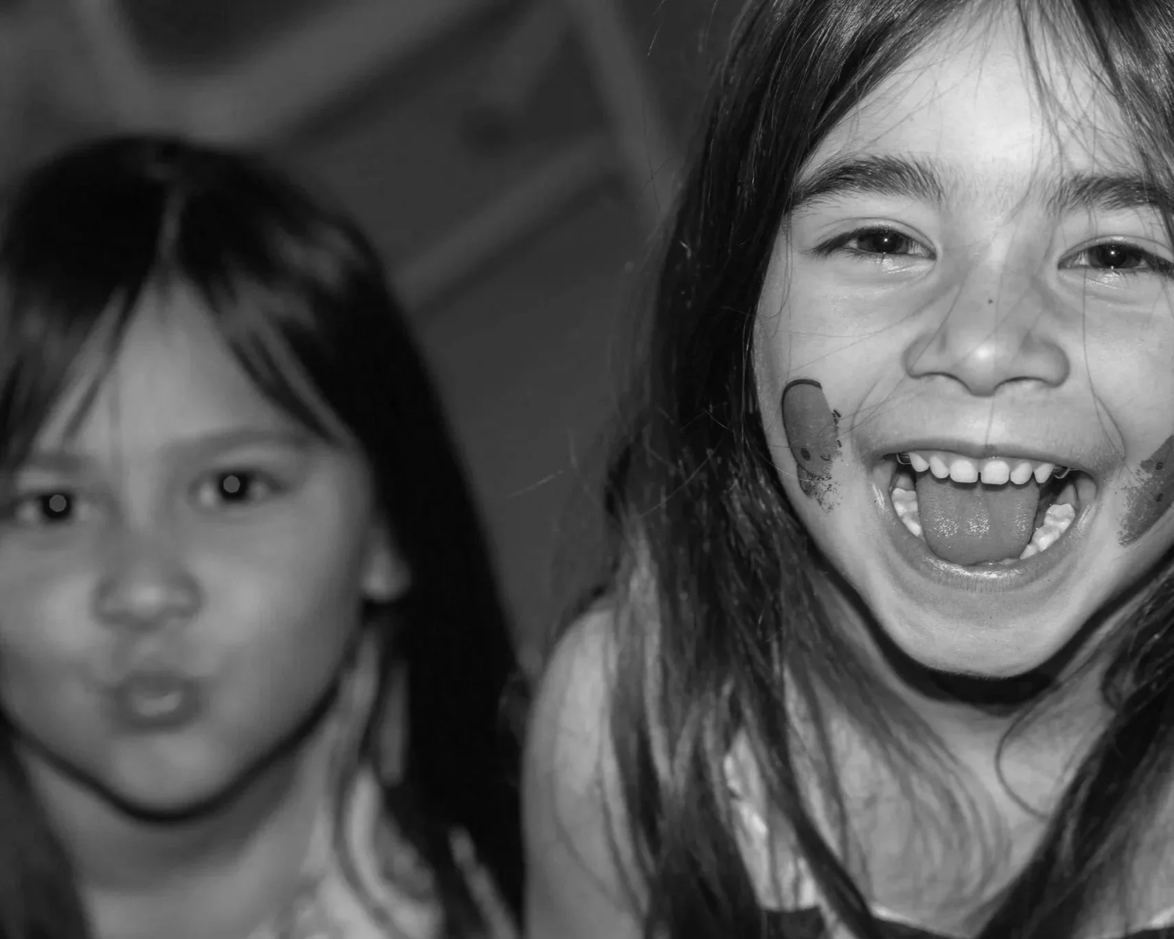 Close-up of two young girls, one smiling with her mouth open, showing her teeth and tongue, with stickers on her face, in black and white.