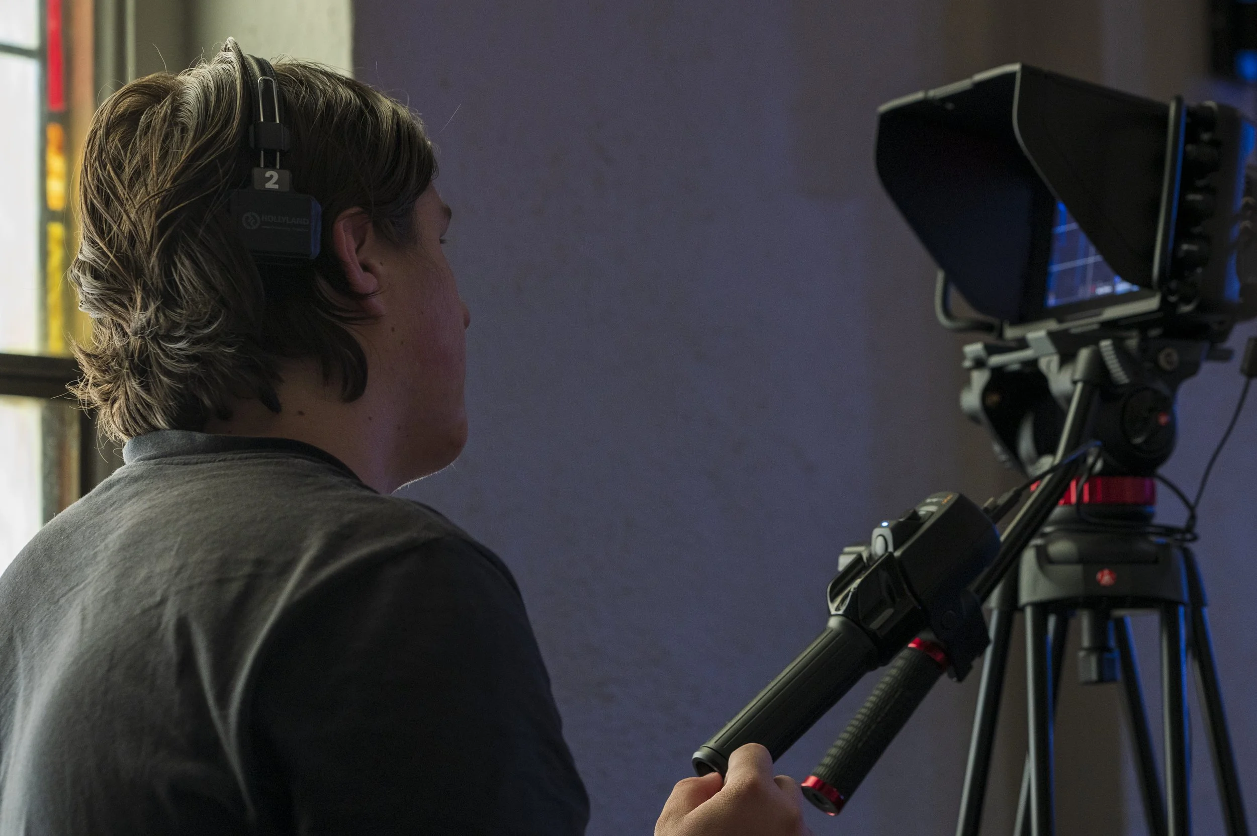 A person with light brown hair, wearing a gray shirt and black headphones, is looking through a professional camera mounted on a tripod.