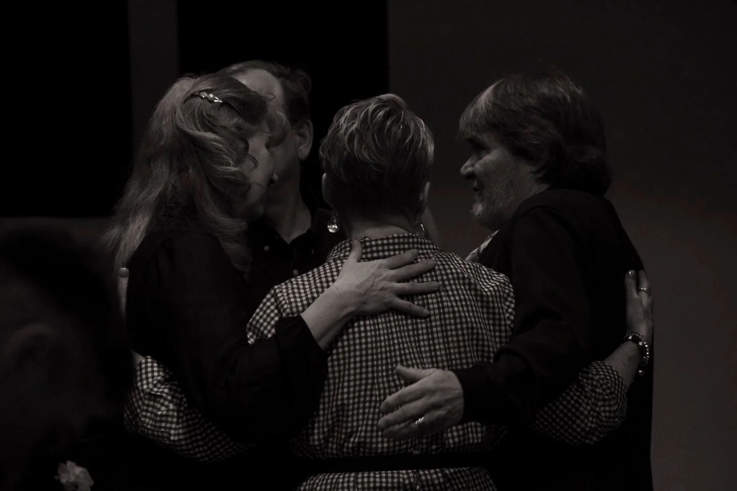 Four people are in a close group hug, touching heads and shoulders, with one person in the center facing away. The scene is in black and white, suggesting an emotional or intimate moment.