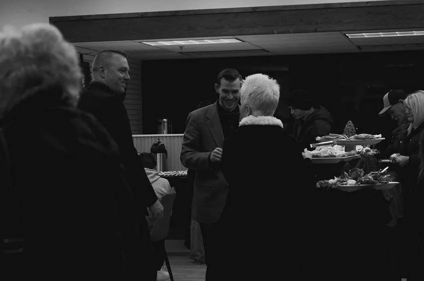 People at a social gathering or event, some engaging in conversation, with food on a table in the background.