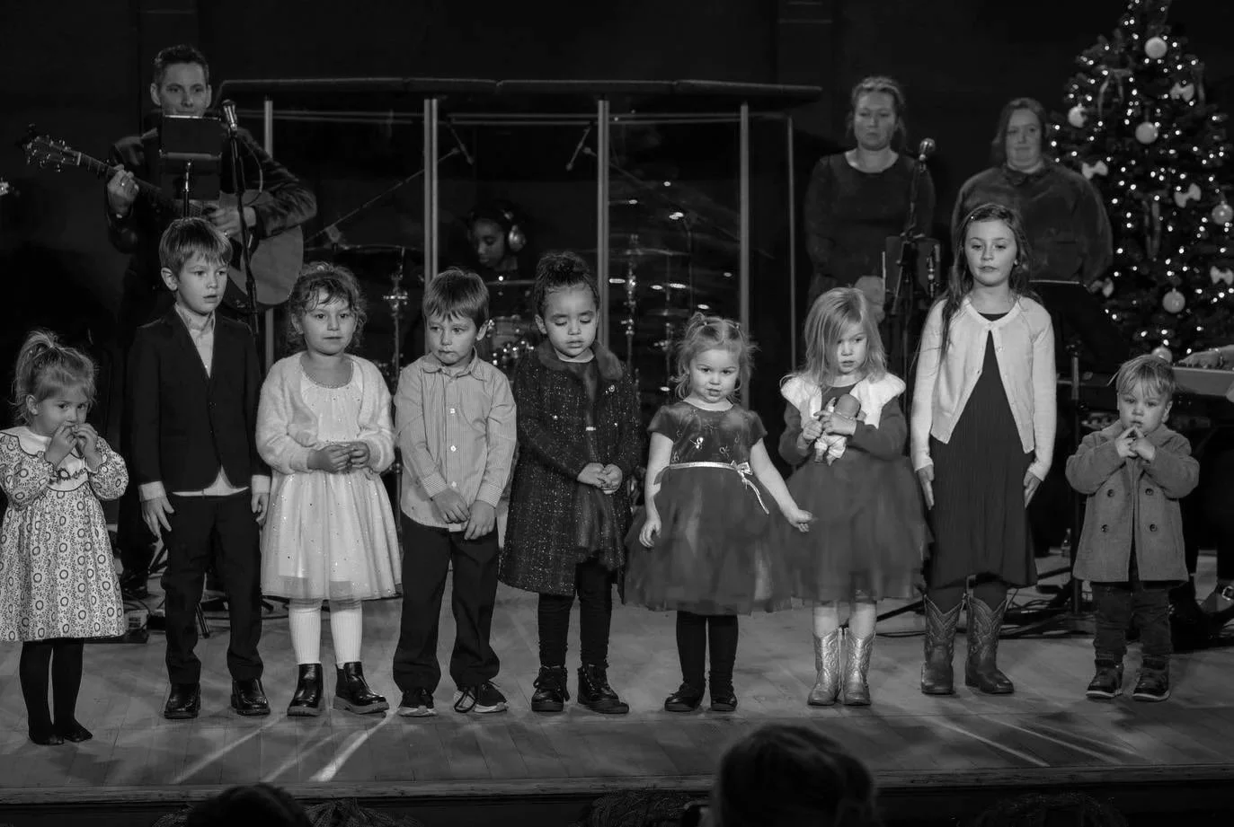 Children performing on stage during a Christmas event, with a decorated Christmas tree in the background, accompanied by musicians.