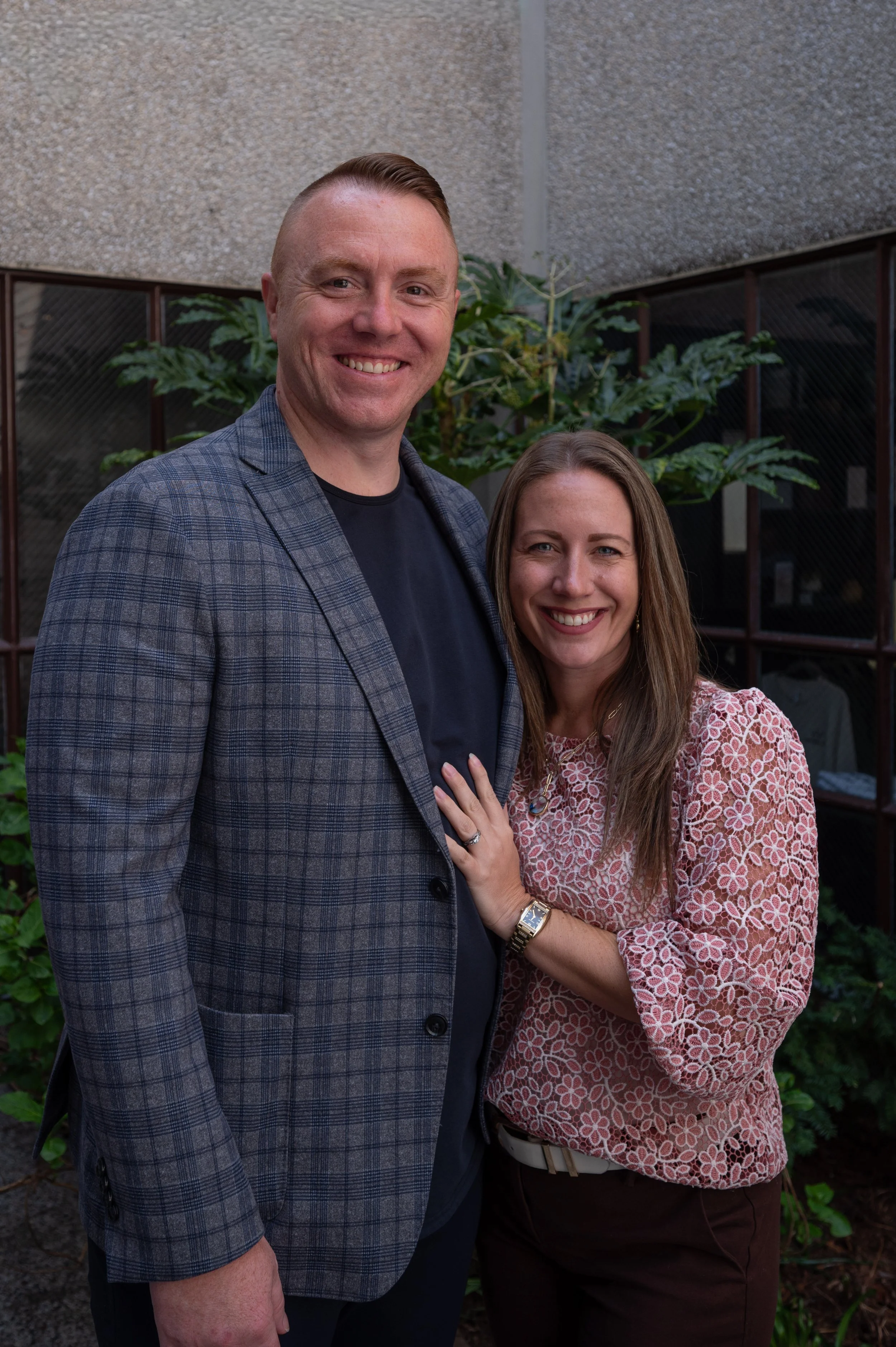 A smiling man and woman standing outdoors in front of greenery and a concrete building wall.