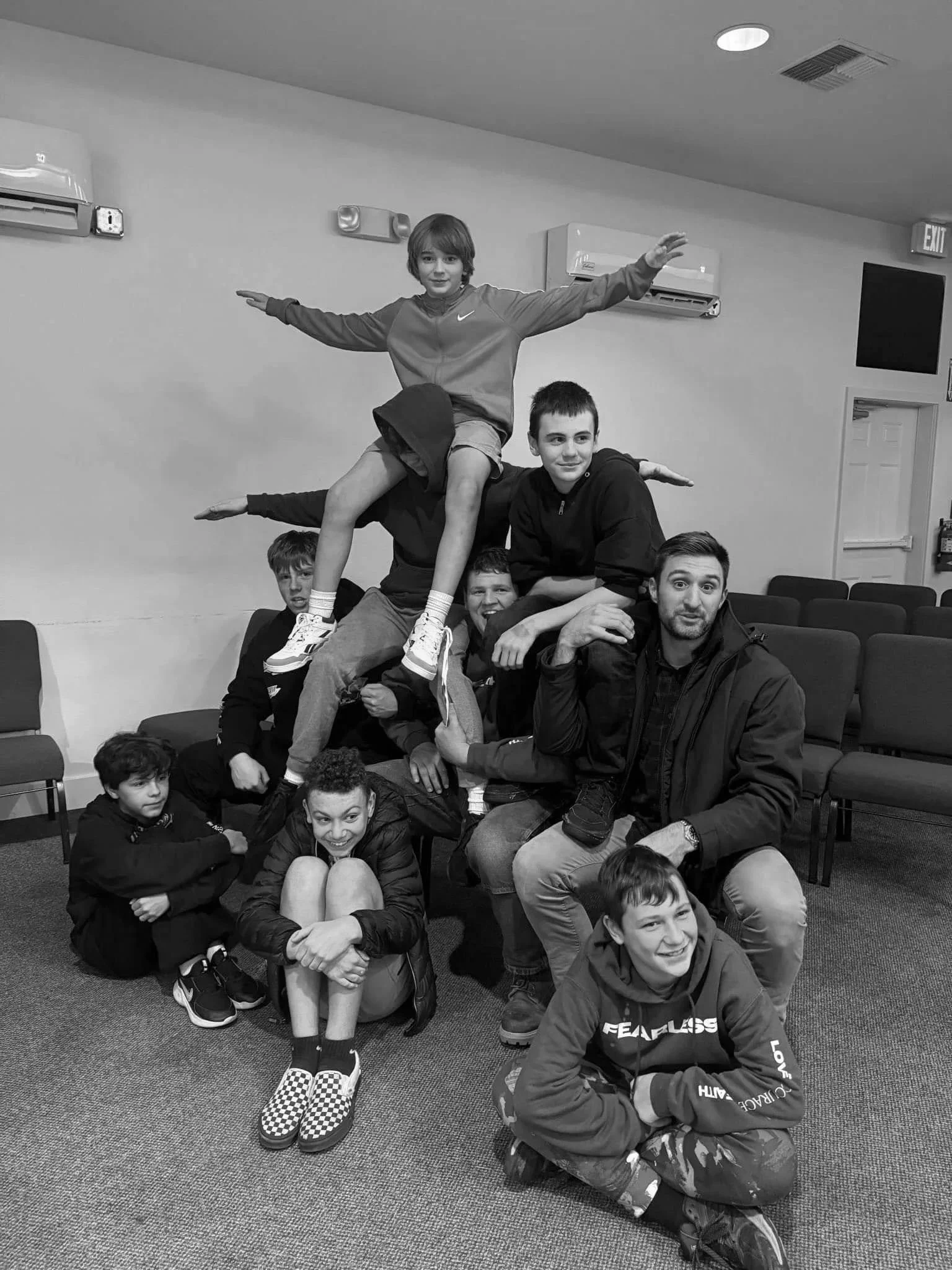 A group of children and adults forming a human pyramid indoors, with a boy at the top extending his arms outward. The people are sitting on chairs and the floor, smiling and looking at the camera.