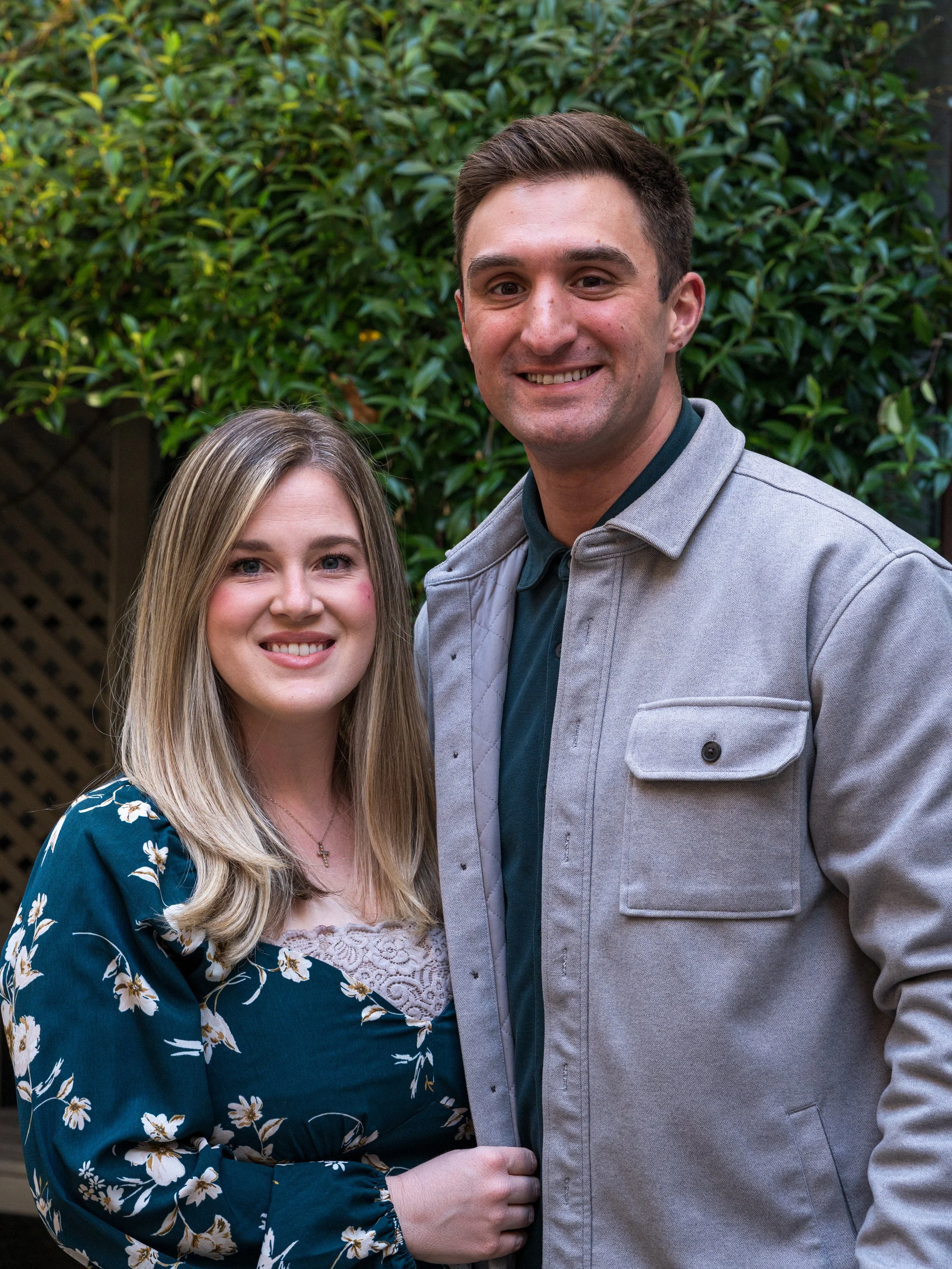 A smiling young woman with blonde hair, wearing a blue floral dress, standing next to a smiling young man with dark hair, wearing a gray jacket and dark shirt. They are outdoors with green bushes in the background.