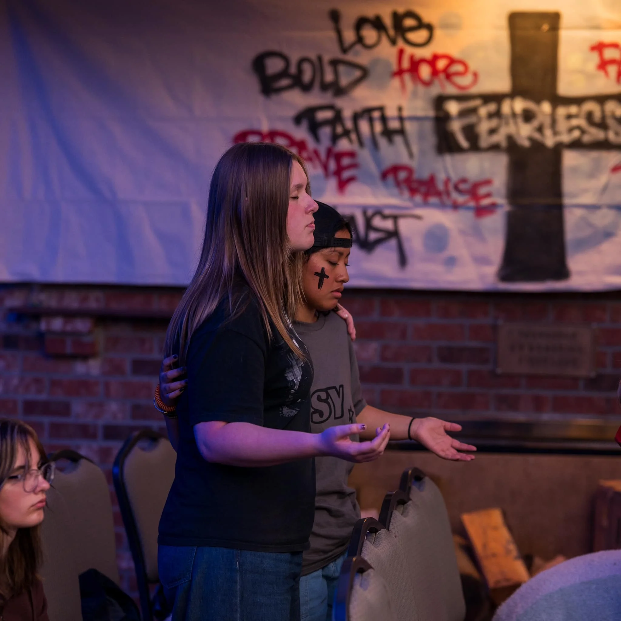 A group of young women standing with their eyes closed, participating in a moment of silence or prayer in a room with graffiti-covered walls, including a prominent cross and words like "Love" and "Peace".