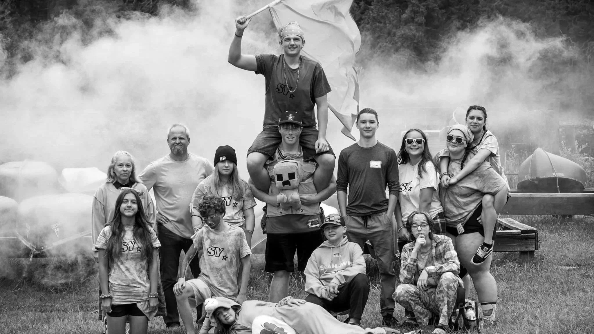 A group of people, mostly children and teenagers, gathered outdoors, with some wearing tie-dye shirts, sunglasses, and hats. One person is standing on the shoulders of another, holding a flag, and there is a smoky background with tents, indicating a festival or outdoor event.