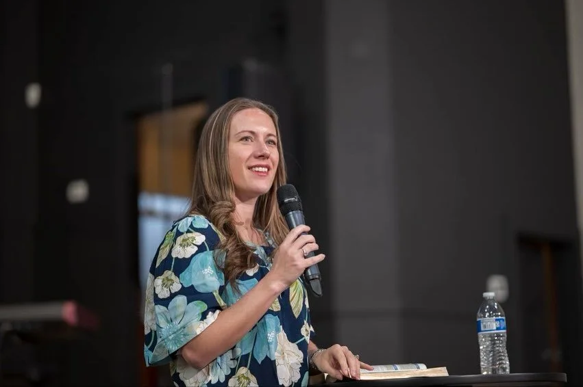 A woman standing at a podium holding a microphone, wearing a floral dress, with a water bottle on the table beside her in an indoor setting.