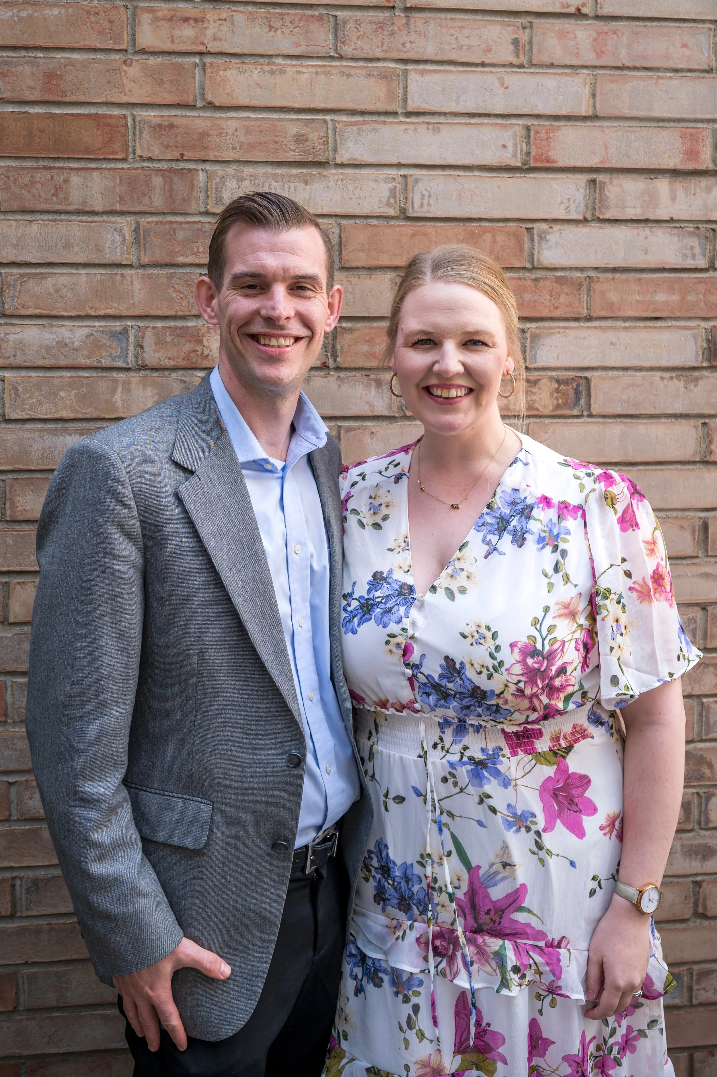 A man and woman smiling and standing together against a brick wall. The man is wearing a gray blazer and blue shirt, and the woman is wearing a floral dress. They are standing close to each other.