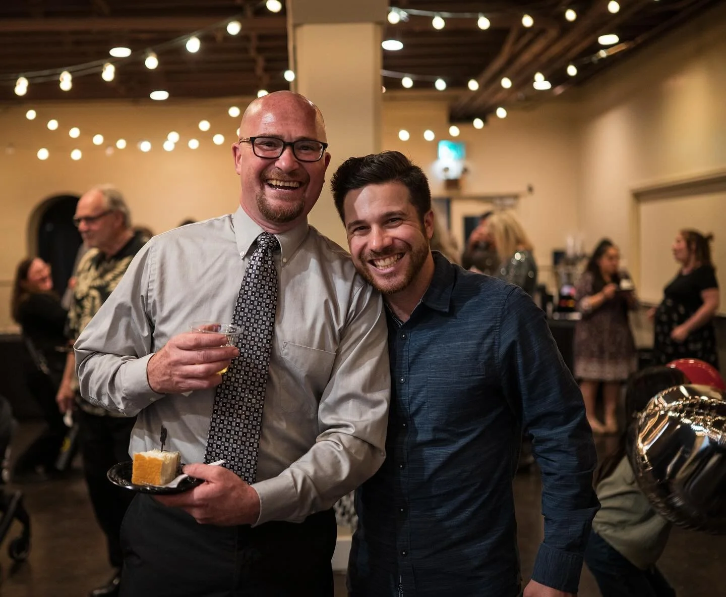 Two smiling men at a celebration, one holding a plate with a slice of cake and a drink, in a decorated indoor venue with string lights and other guests in the background.