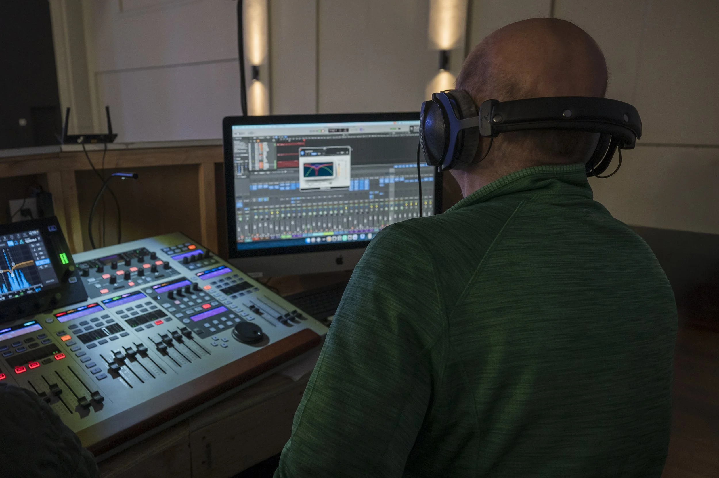 A person wearing headphones working at a digital audio workstation with a computer screen displaying music production software, and mixing equipment with colorful controls in a dimly lit room.