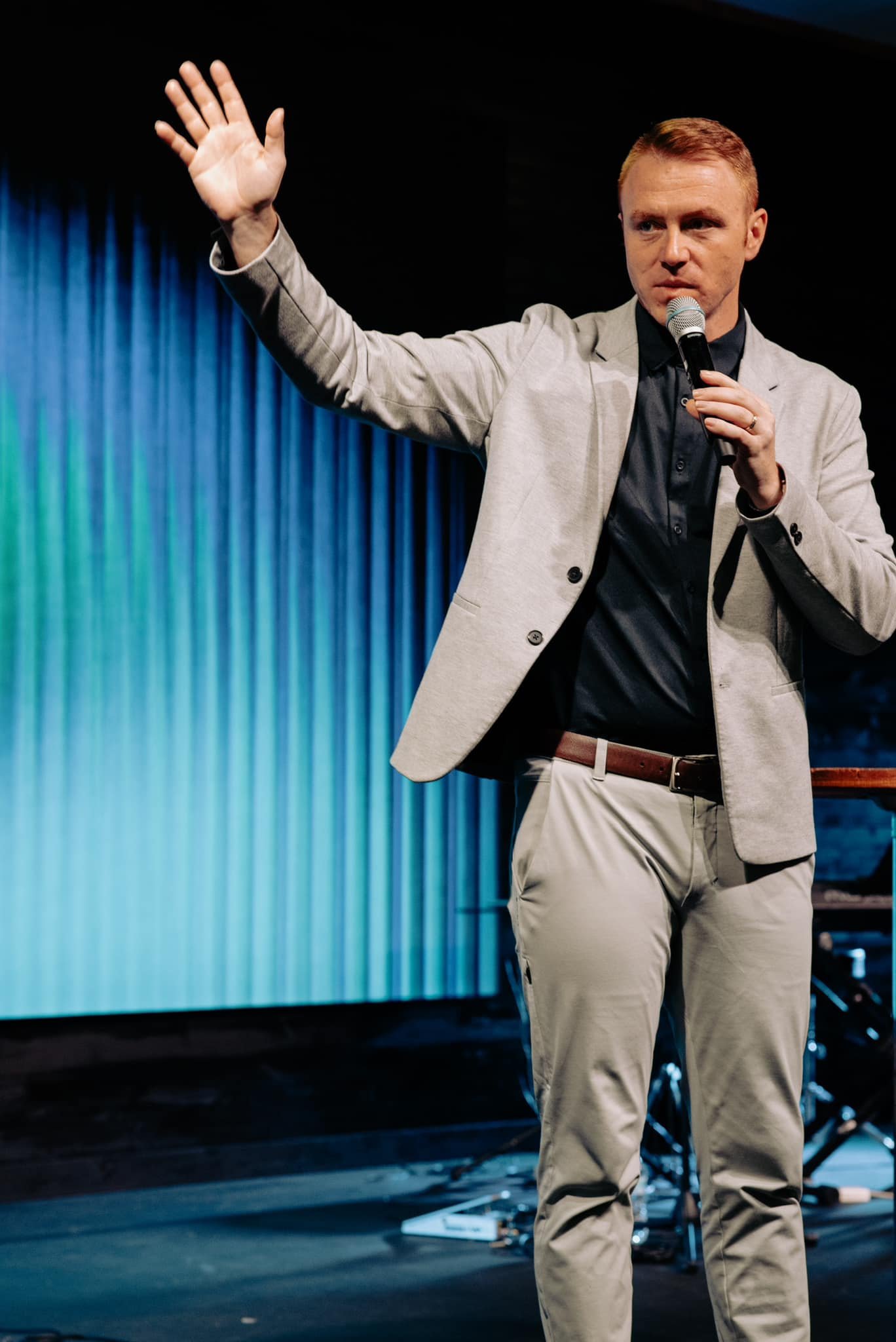 A man with short red hair wearing a light gray blazer, black shirt, and beige pants, holding a microphone, gestures with his left hand while standing in front of a blue curtain on a stage.
