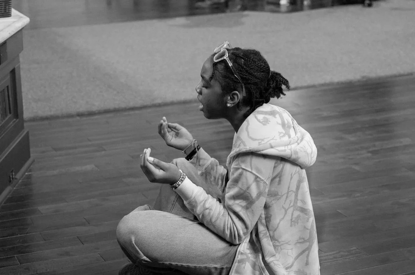 A young person with glasses on their head, sitting cross-legged on a wooden floor, appears to be praying or meditating with closed eyes and hands held up.