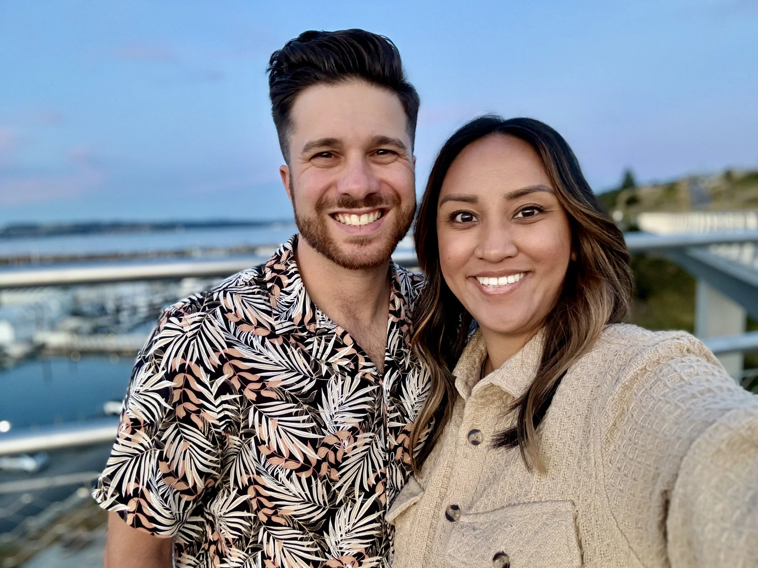 A smiling man and woman taking a selfie outdoors, with a body of water and a bridge in the background during sunset.