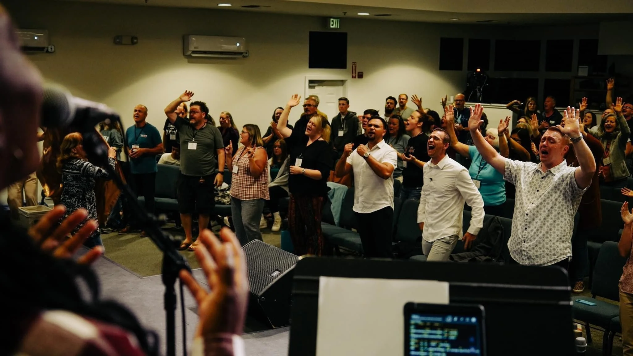 A group of people at a conference or event standing with hands raised, some singing or cheering, in an indoor venue with a stage, microphone, and camera.