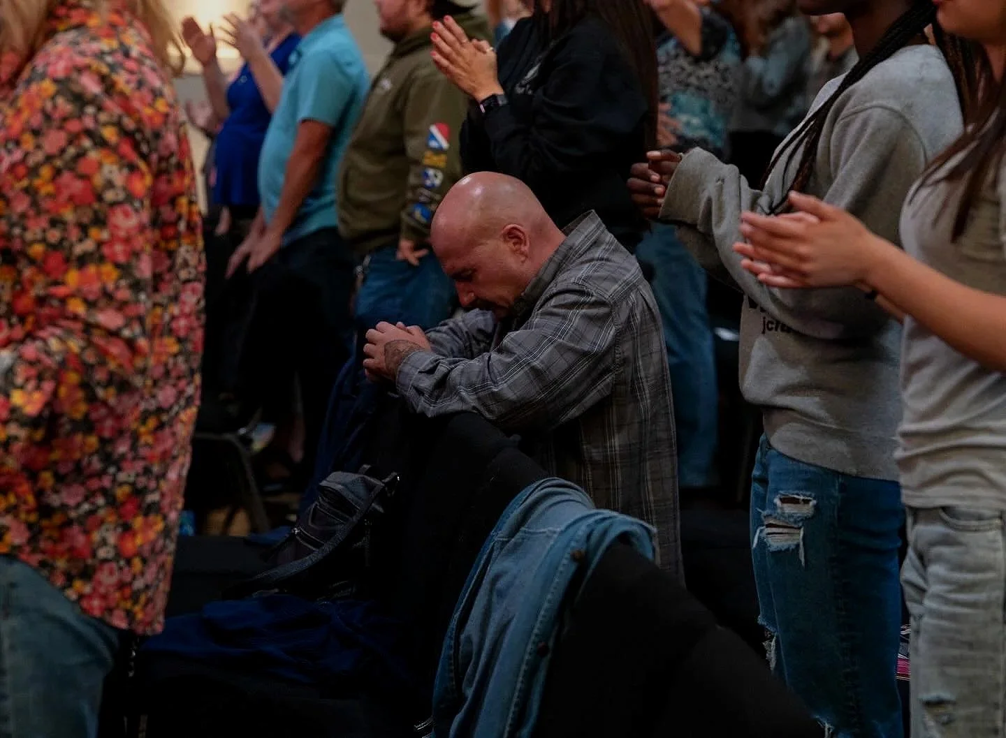 People praying, with a man in a plaid shirt kneeling and bowing his head, surrounded by others with hands clasped or raised.