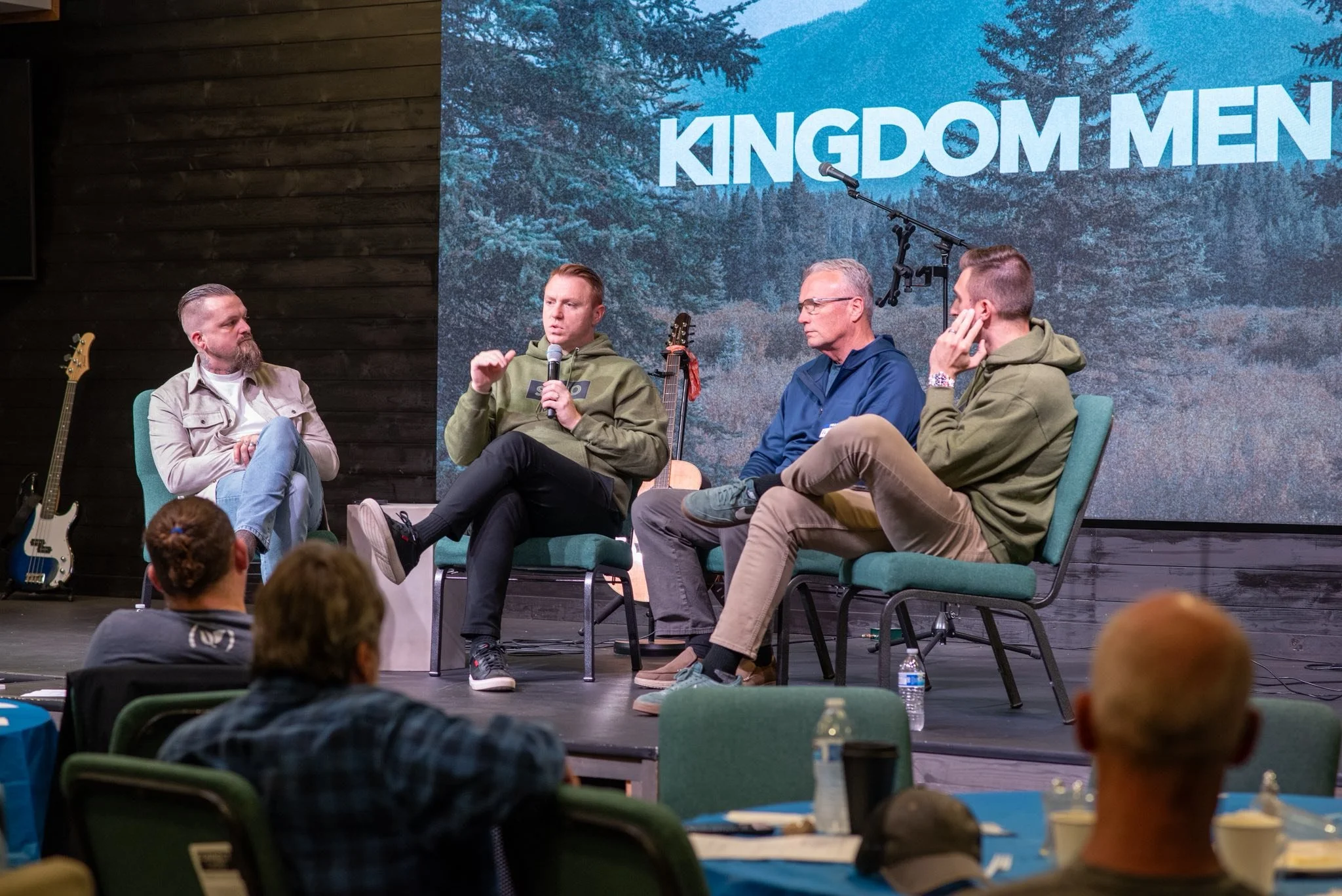 A panel discussion on stage at an event called 'Kingdom Men'. Four men are seated on chairs, two of them are speaking into microphones. An audience is watching, and the background features a digital screen displaying a mountain forest scene and the text 'KINGDOM MEN'.