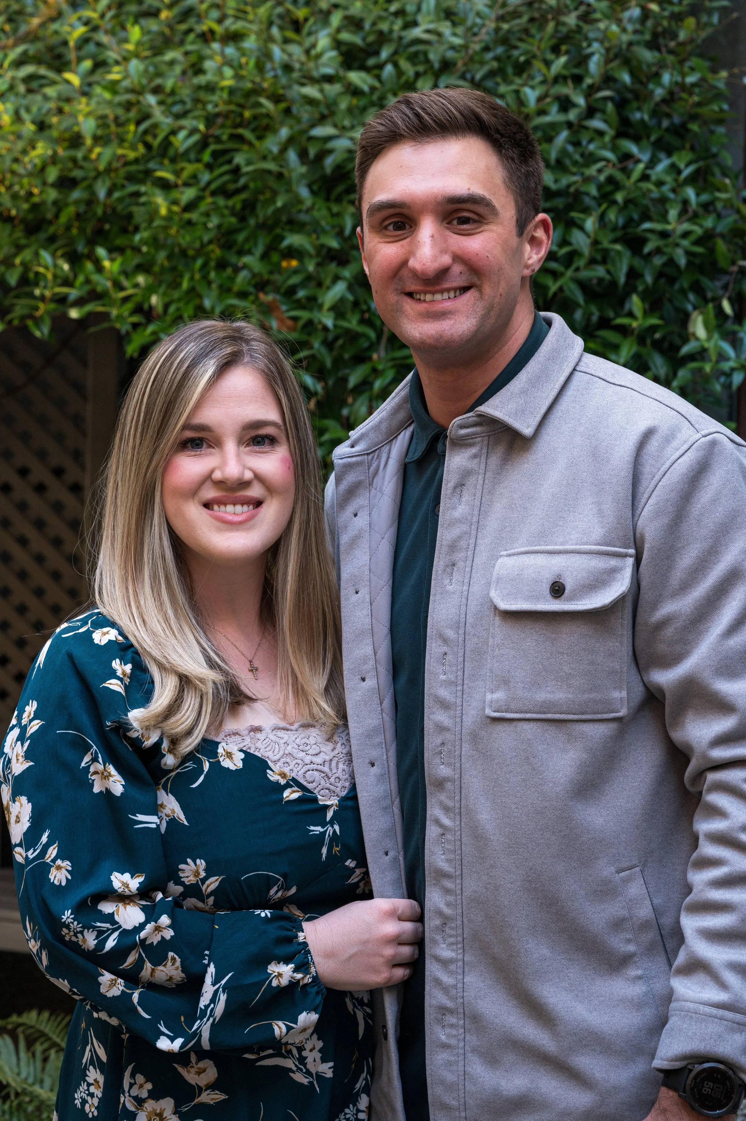 A young woman with long blonde hair, smiling, wearing a dark teal dress with white and yellow floral pattern, standing outdoors beside a young man with short dark hair, smiling, wearing a light gray jacket over a dark shirt, in front of green leafy bushes.