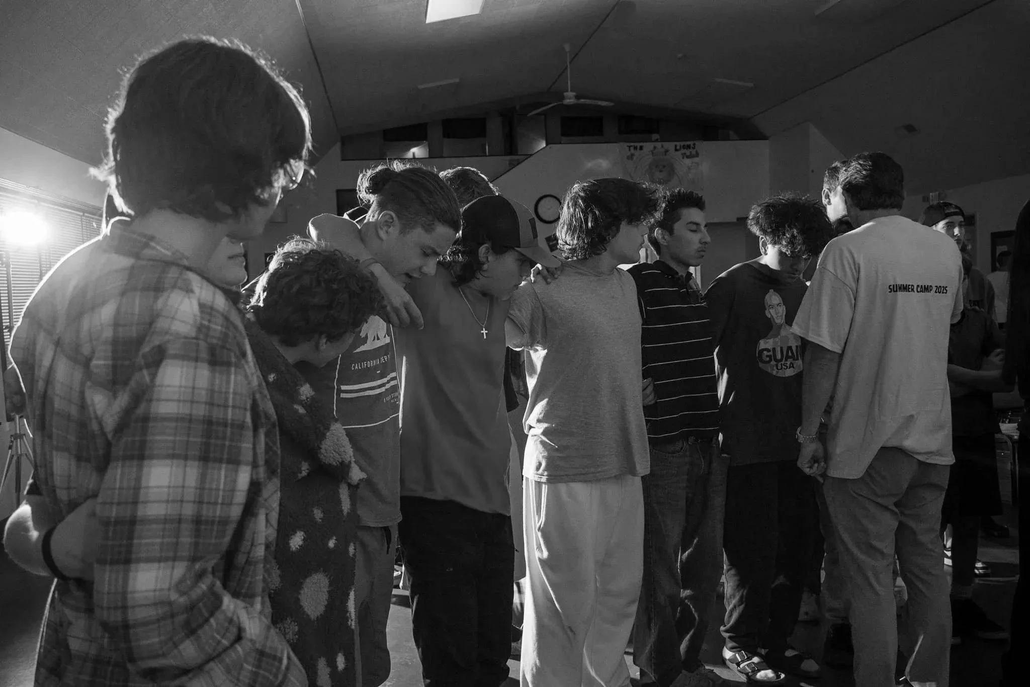 A group of teenagers standing shoulder to shoulder with heads bowed and eyes closed, gathered indoors with subdued lighting.
